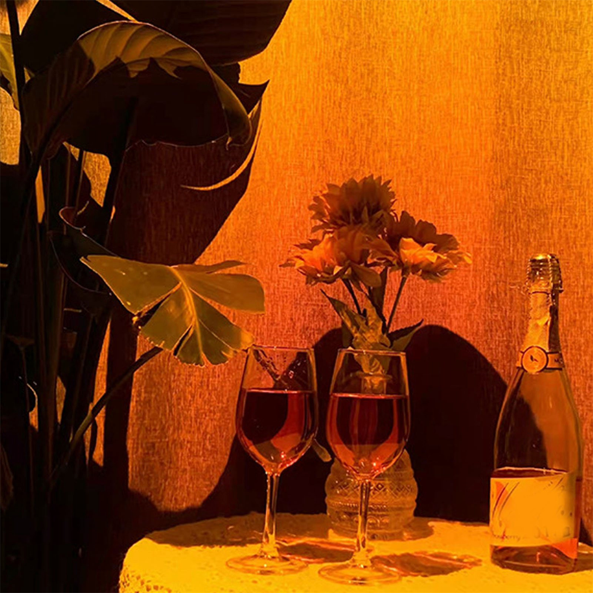 Ein Tisch mit Weingläsern und einer Flasche. Links ist eine Pflanze, und die Szene hat einen orangen Schein.
