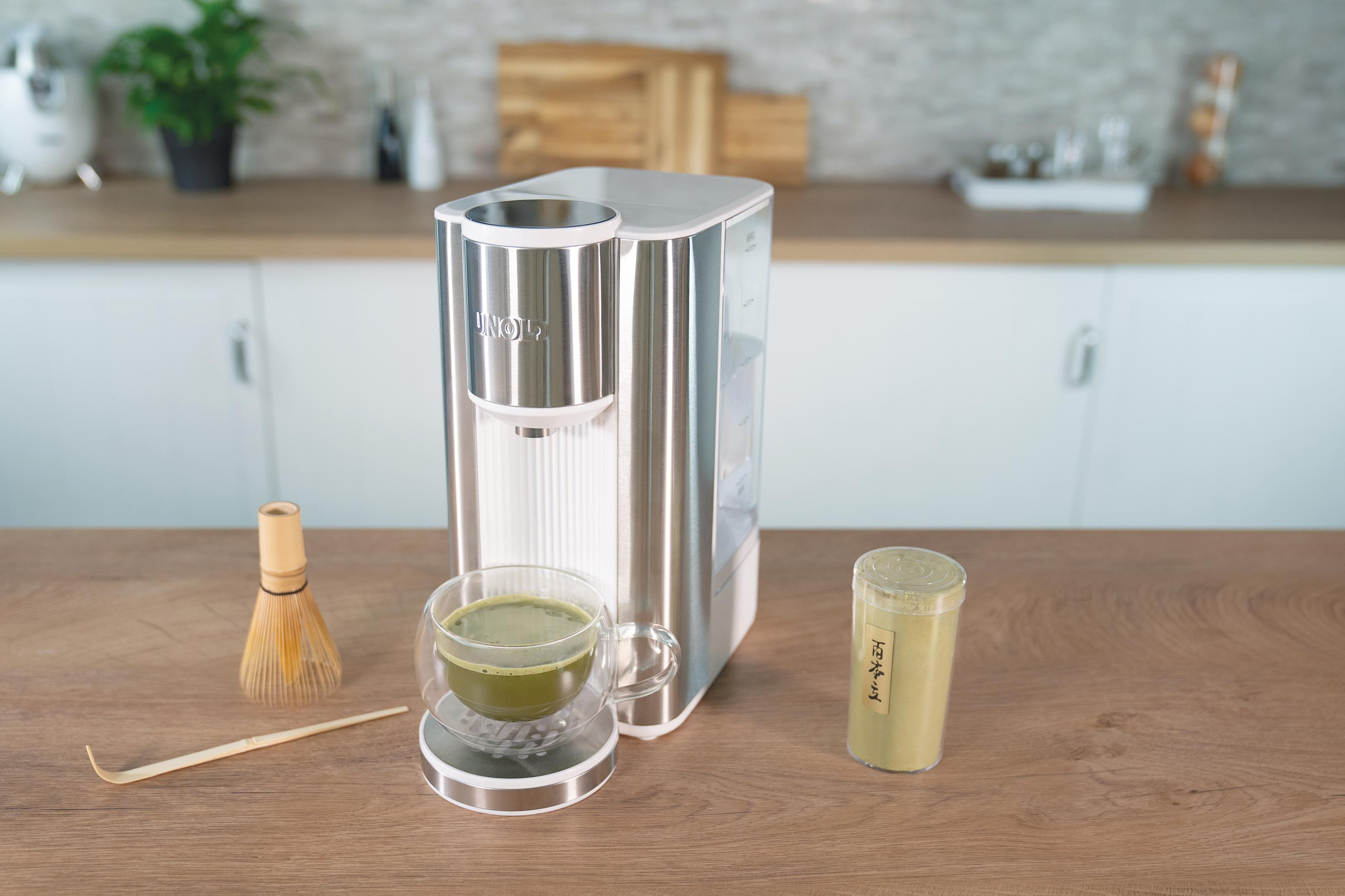 Ein moderner silberner Wasserspender mit einer Tasse Matcha-Tee und Zutaten auf einer Holzoberfläche.