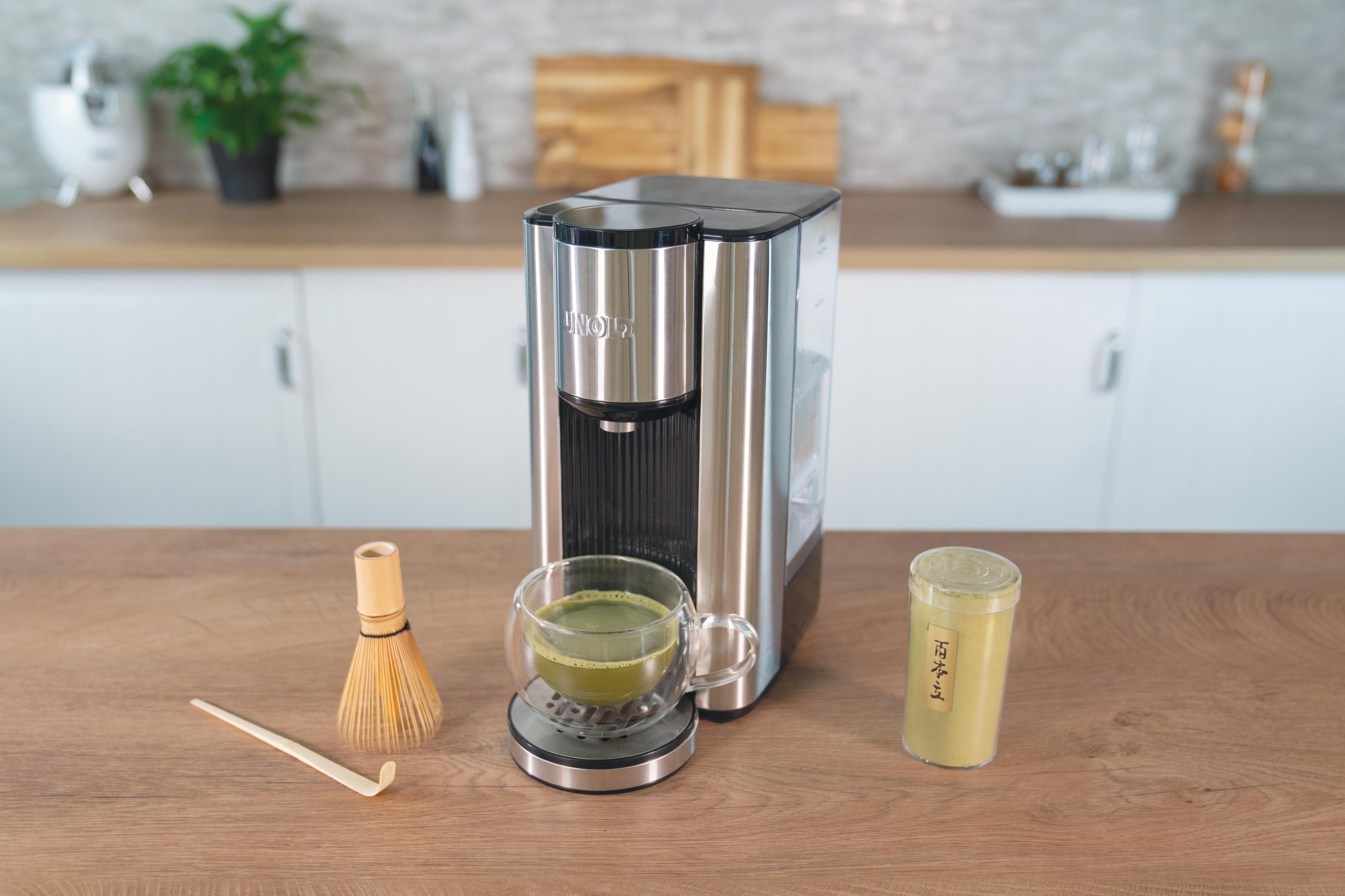 Eine silberne und schwarze Matcha-Maschine mit Matcha in einem Glas und Pulver daneben.