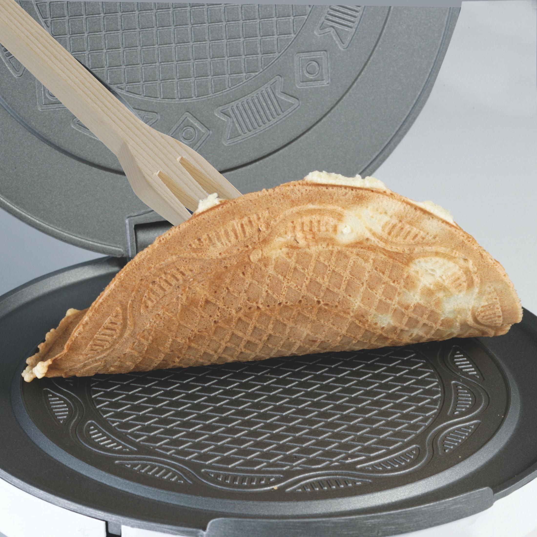 Waffeleisen mit Waffel. Die Waffel ist hellbraun und hat eine Textur. Holzgabel liegt daneben.