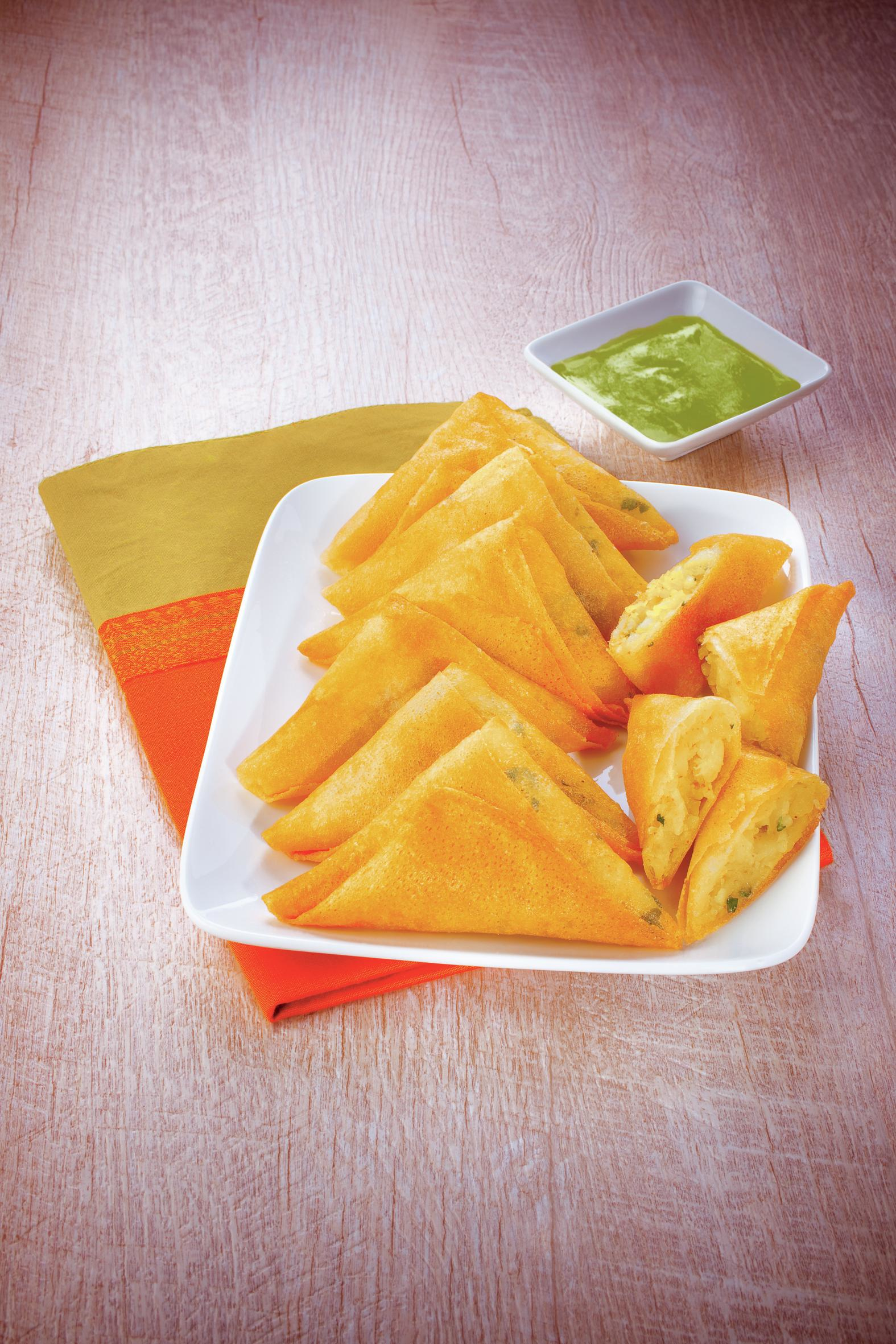 Talerz złotych samosów z zielonym chutneyem w białym naczyniu na drewnianym stole z pomarańczową i żółtą serwetą.