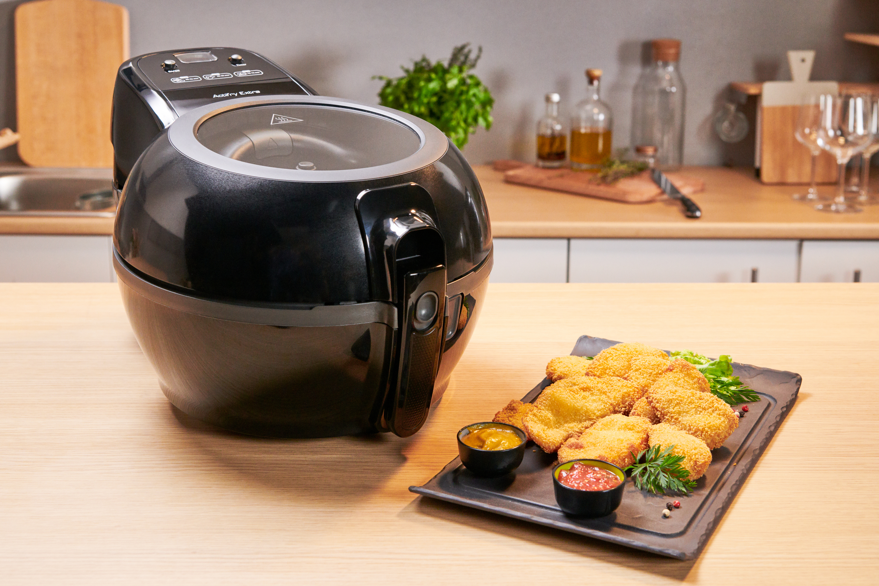 Una freidora de aire con comida frita en un plato y salsas para mojar, sobre una mesa de madera con ambiente de cocina.