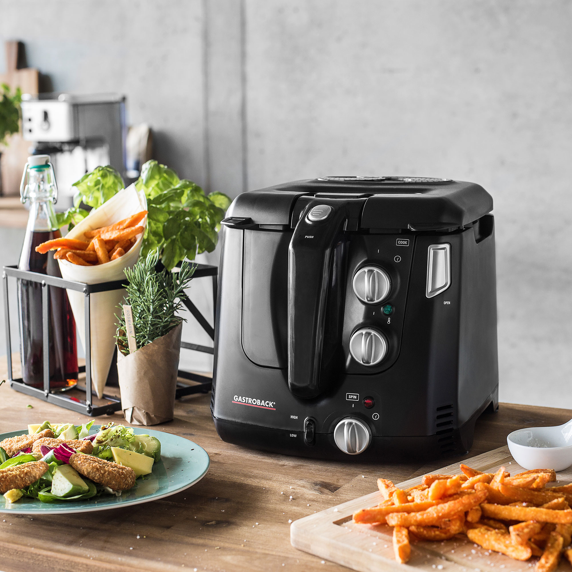 Czarna frytkownica z frytkami i innymi produktami spożywczymi, ustawiona na drewnianym stole w kuchni.