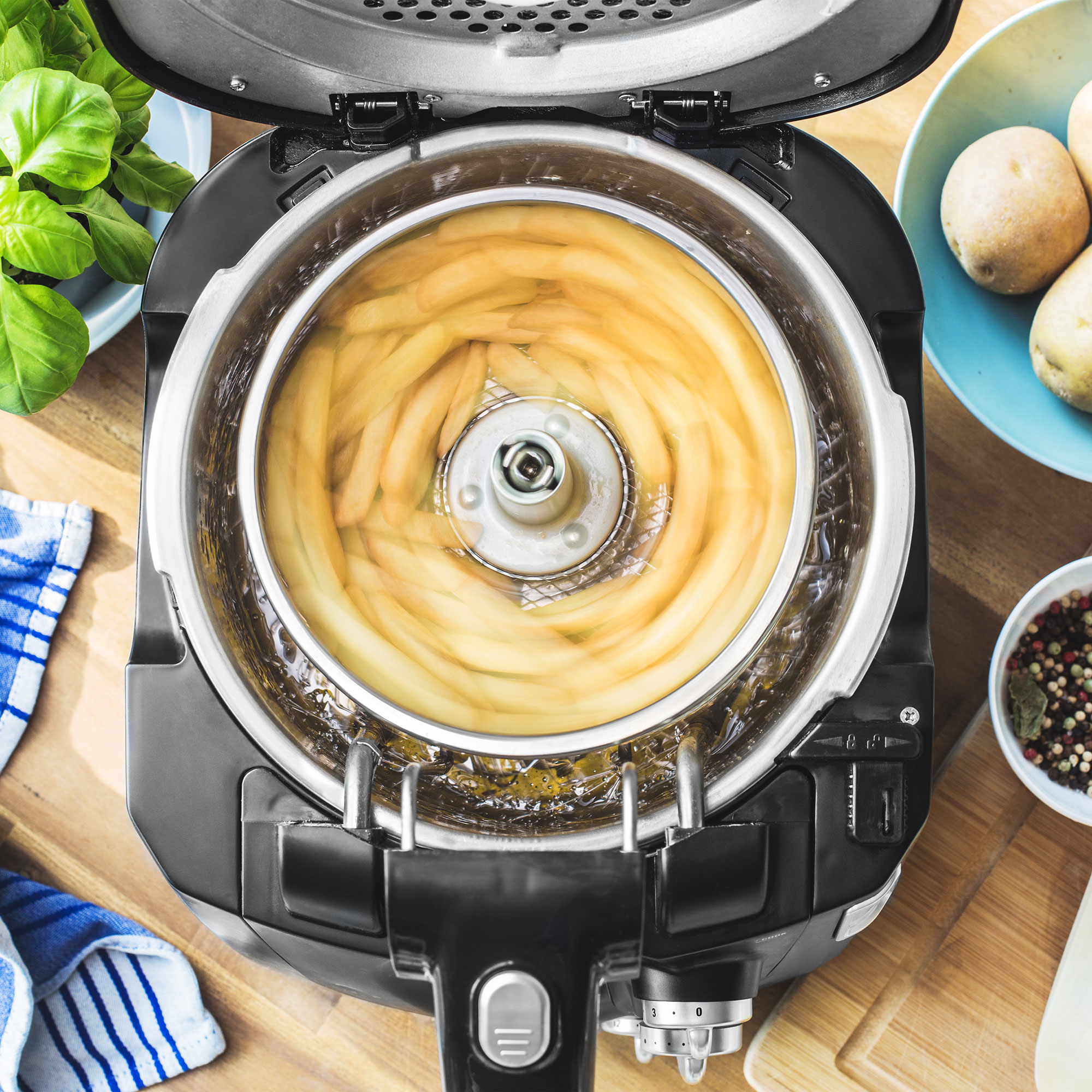 Frytownica powietrzna z frytkami w środku. Ziemniaki i bazylia w tle.