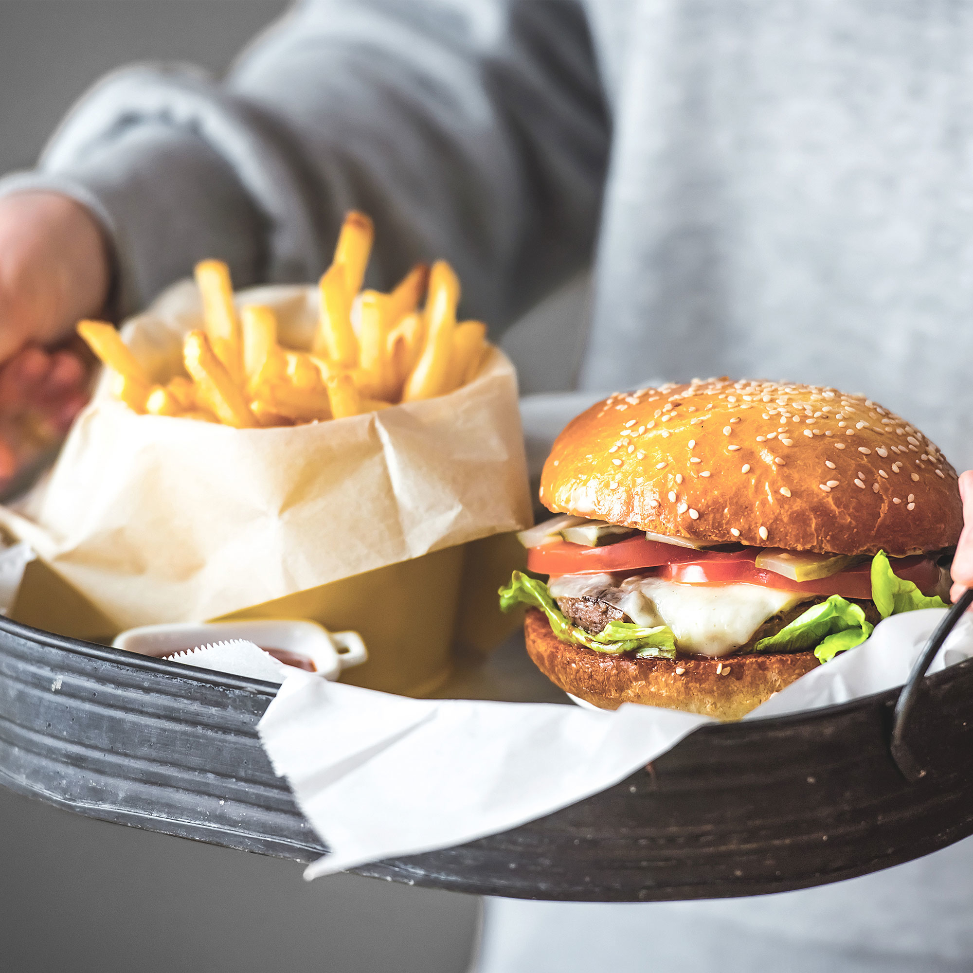 Osoba trzymająca tacę z burgerem i frytkami, z małym pojemnikiem na ketchup.