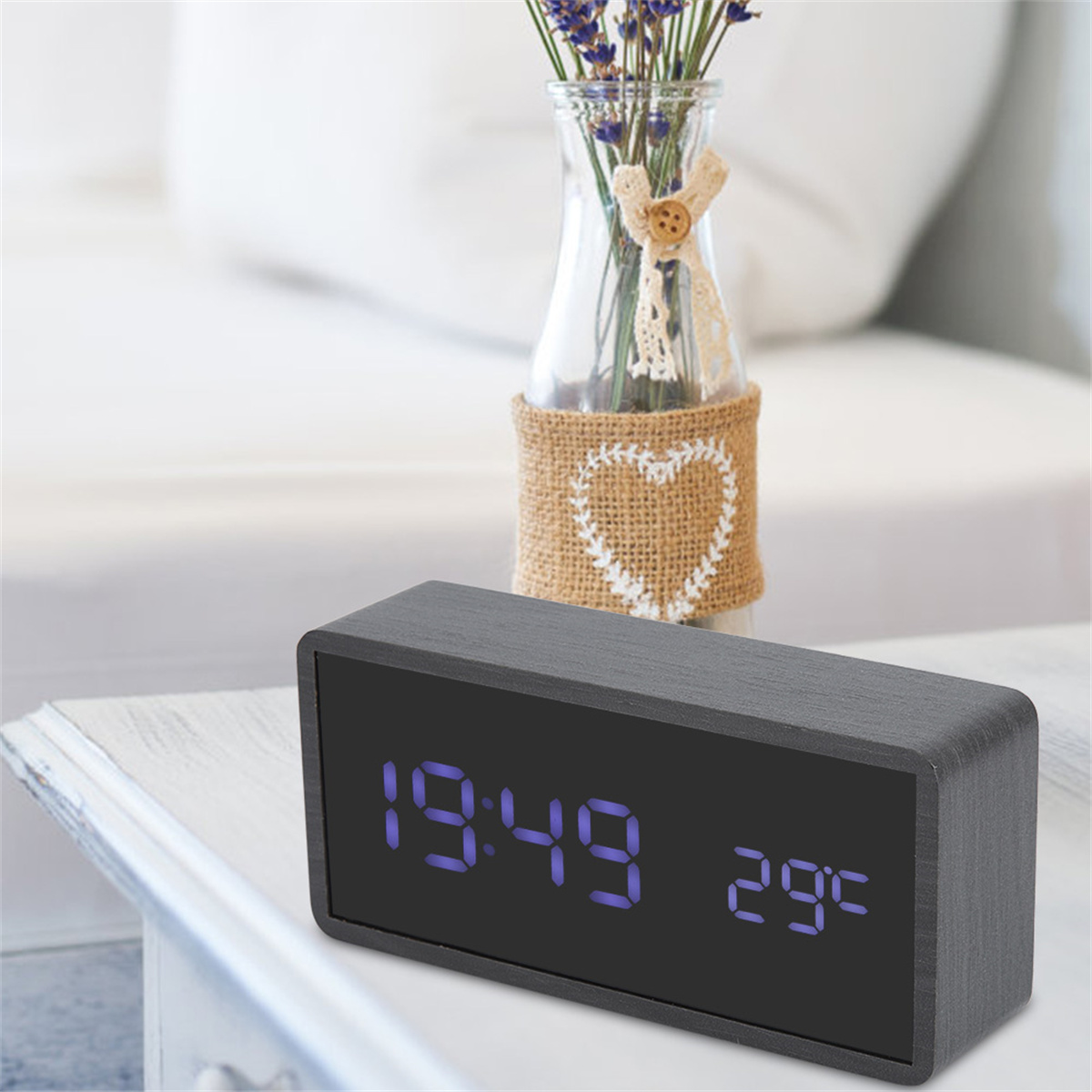 Eine graue Digitaluhr zeigt 19:49 und 29°C an. Eine Vase mit Blumen steht dahinter.