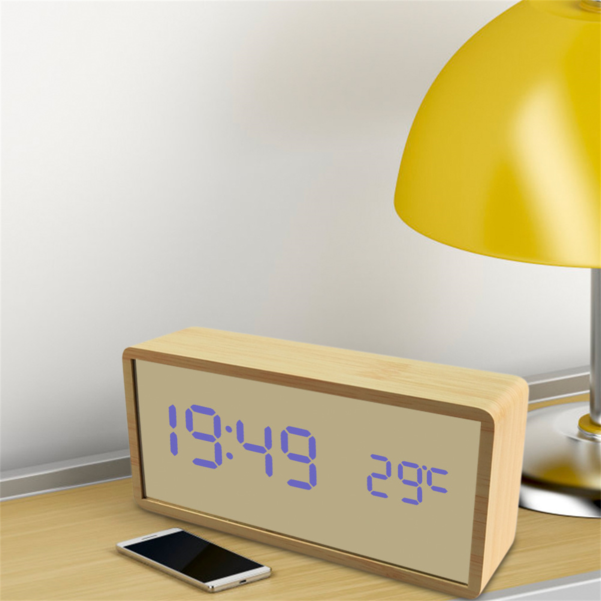 Ein digitaler Wecker aus Holz, der 19:49 und 29°C anzeigt. Auf dem Tisch liegt ein Telefon. Gelbe Lampe in der Nähe.