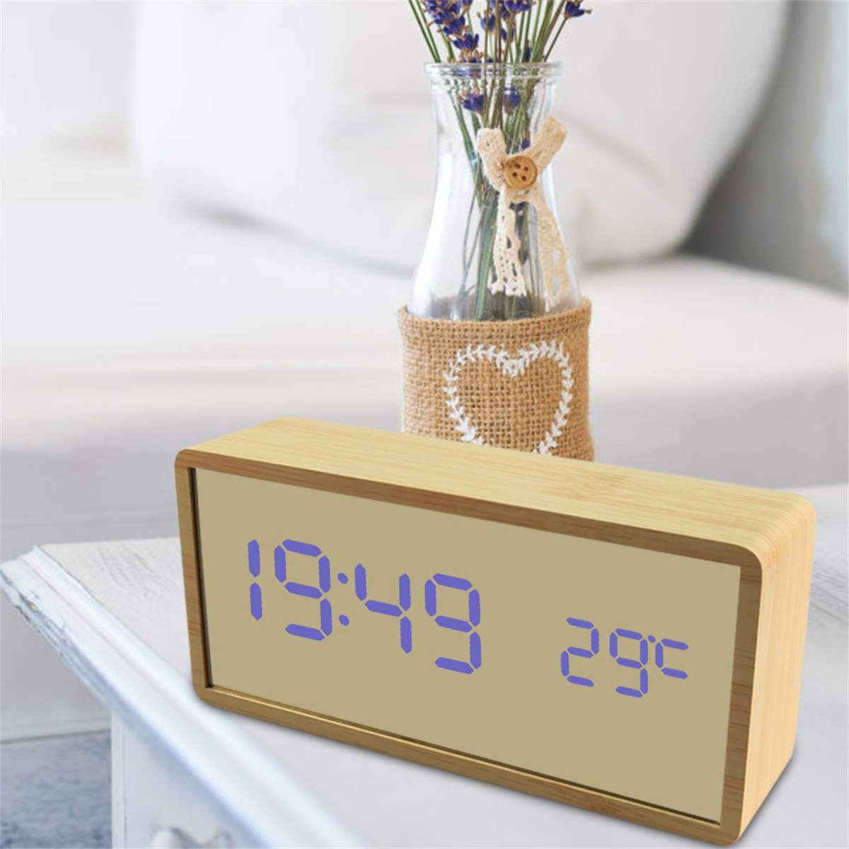 Ein Nachttischuhr aus Holz zeigt 19:49 und 29°C an. Sie steht auf einem Tisch mit einer Blumenvase im Hintergrund.
