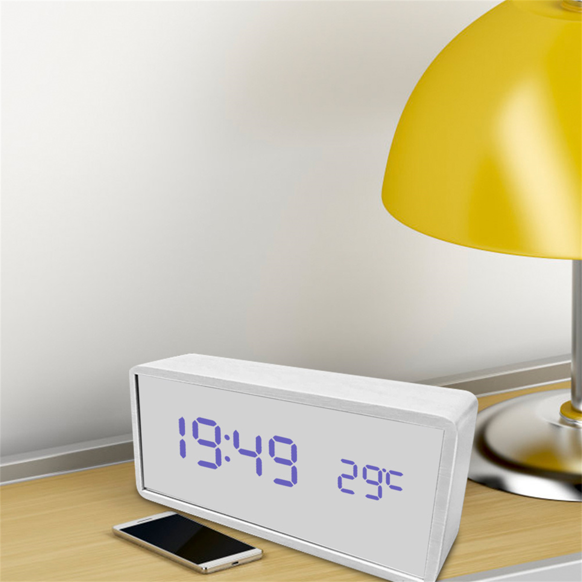 Digitaluhr mit 19:49 und 29C. Smartphone auf dem Tisch, gelbe Lampe, weißer Wandhintergrund.