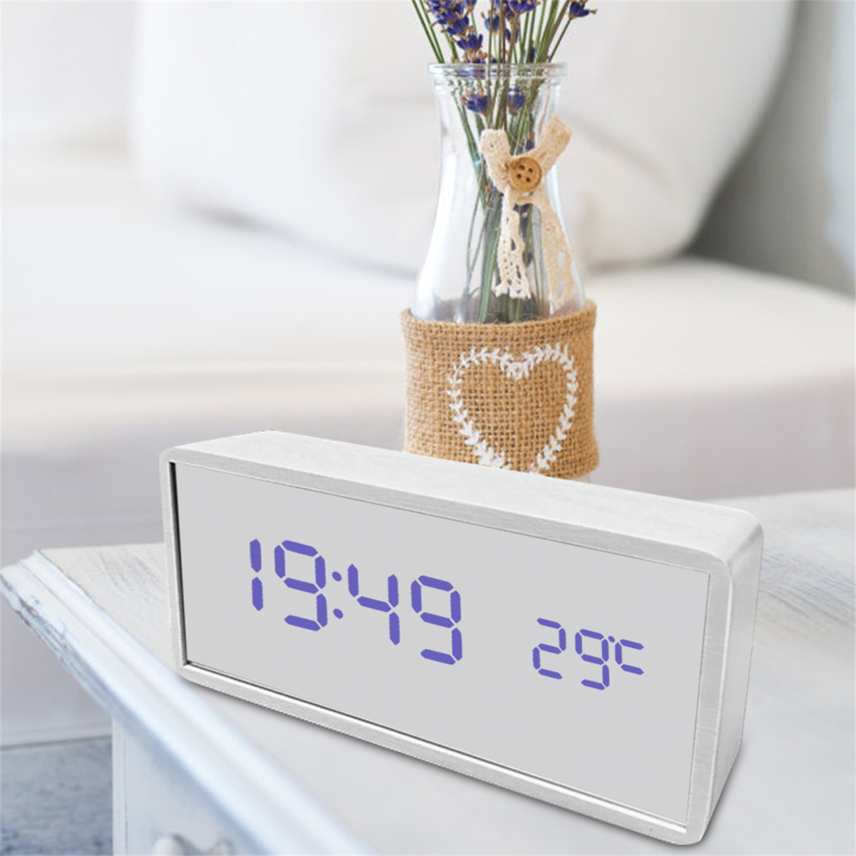 Digitaluhr mit 19:49 und 29°C auf weißer Oberfläche. Blumenvase im Hintergrund, Bett unscharf.
