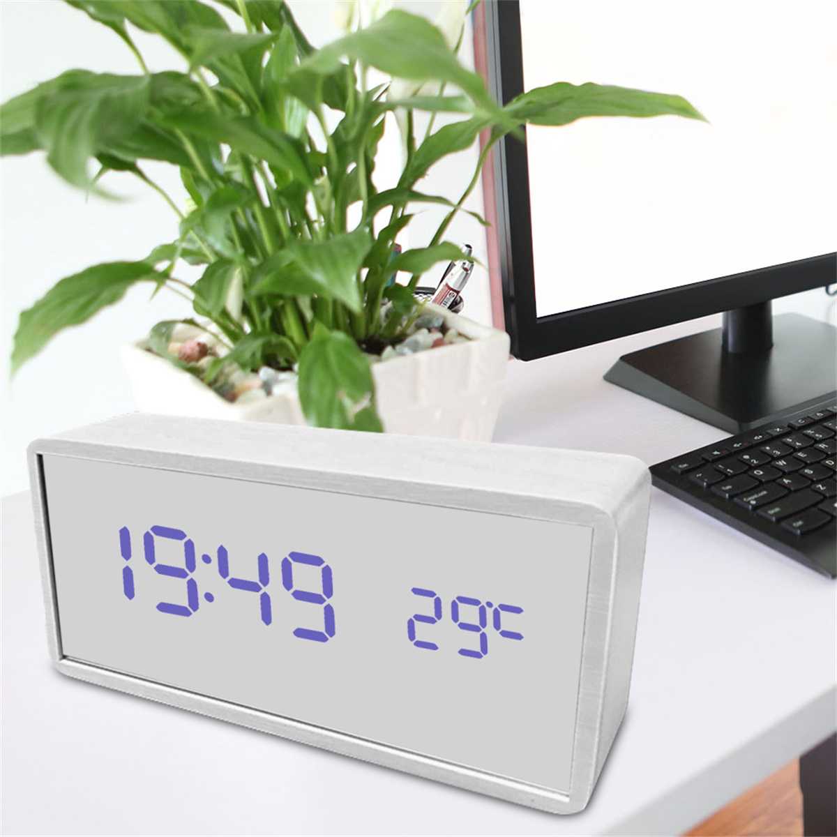 Eine weiße Digitaluhr zeigt 19:49 und 29°C an. Eine Pflanze und ein Computer befinden sich im Hintergrund.