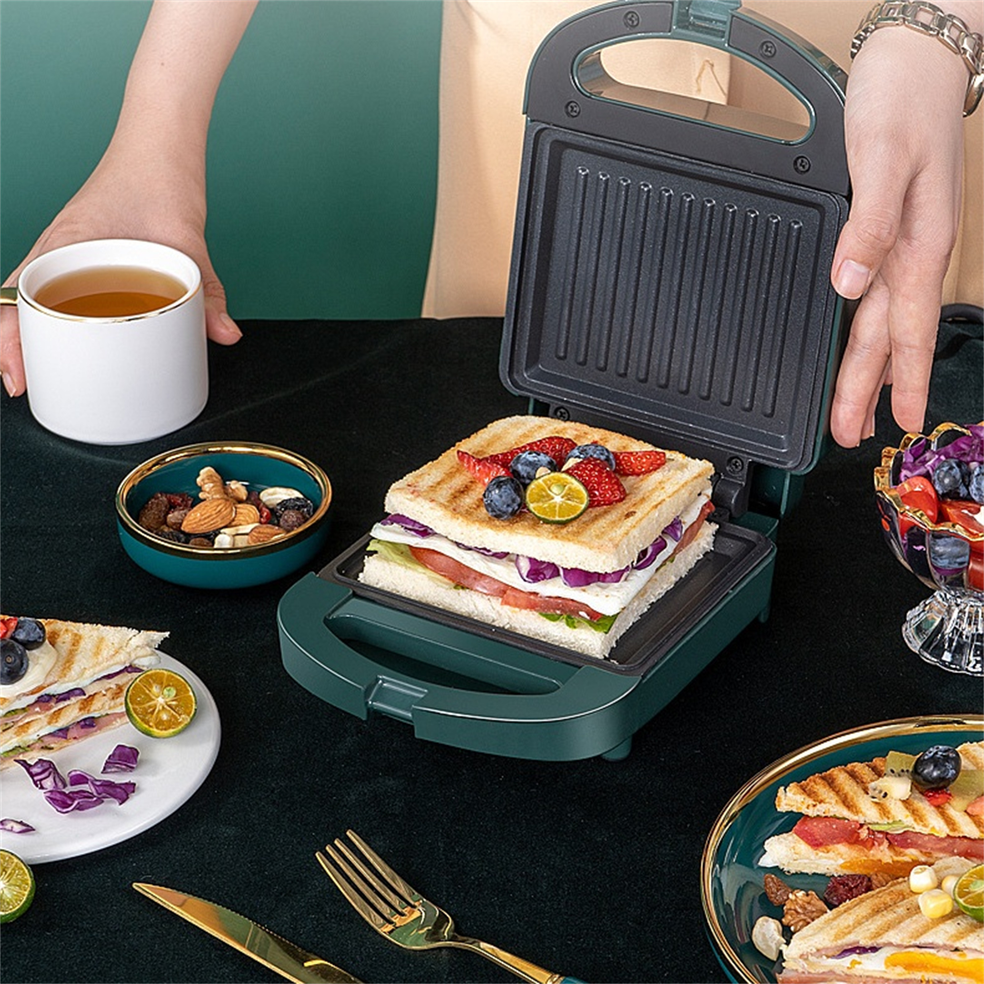 Sandwichmaker mit einem fertigen Sandwich. Mehrere Beilagen stehen auf einem dunklen Tisch.