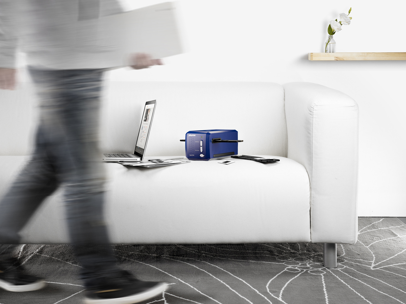 Osoba przechodzi obok białej kanapy ze skanerem, laptopem i zdjęciami. W tle roślina i półka.