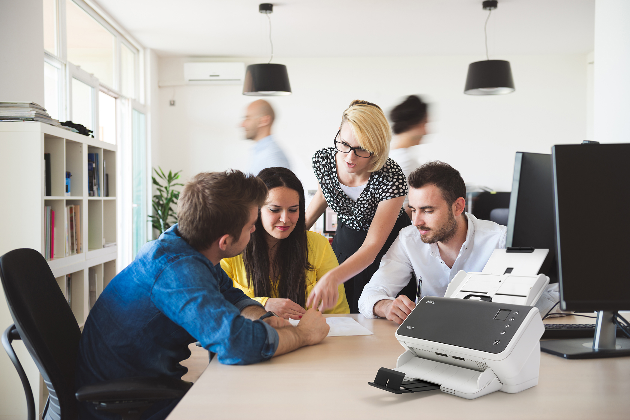 Scena biurowa z ludźmi wokół stołu. Jedna osoba wskazuje na skaner.
