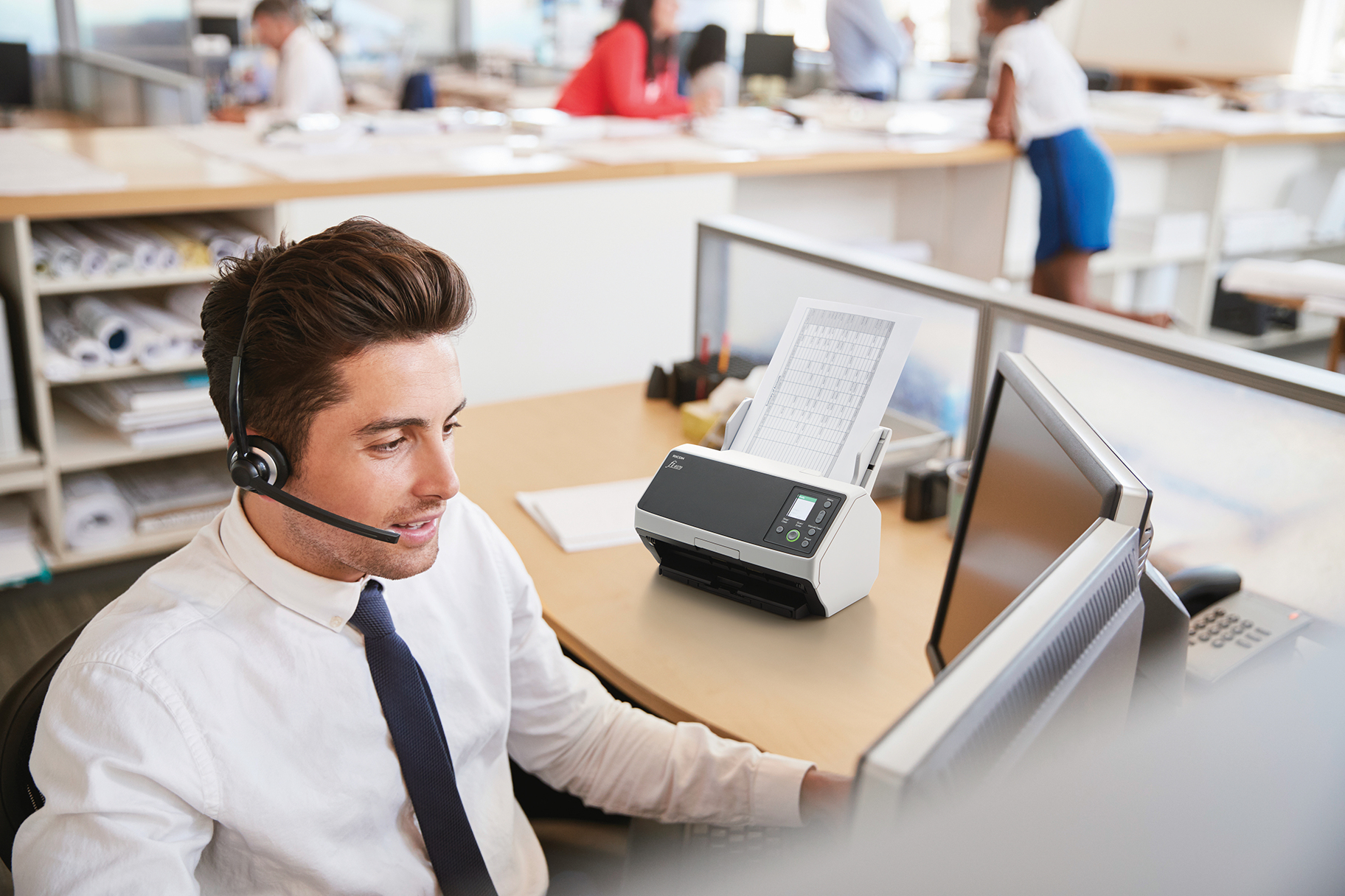 Mężczyzna w zestawie słuchawkowym, pracujący przy biurku ze skanerem i komputerem.
