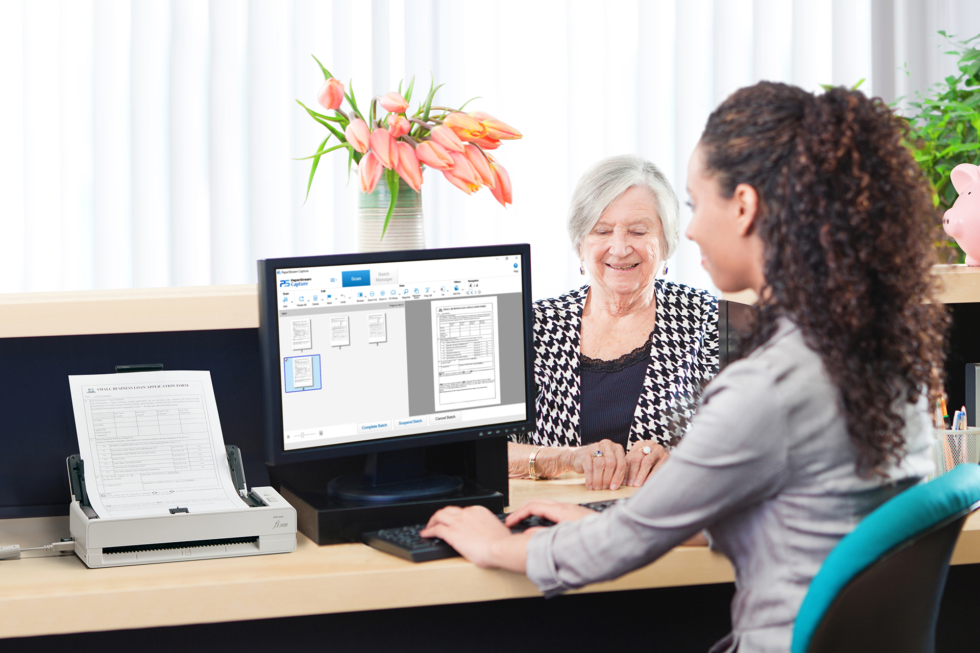 Kobieta przy biurku pomaga starszej pani. Komputer wyświetla zeskanowany dokument. W tle tulipany.