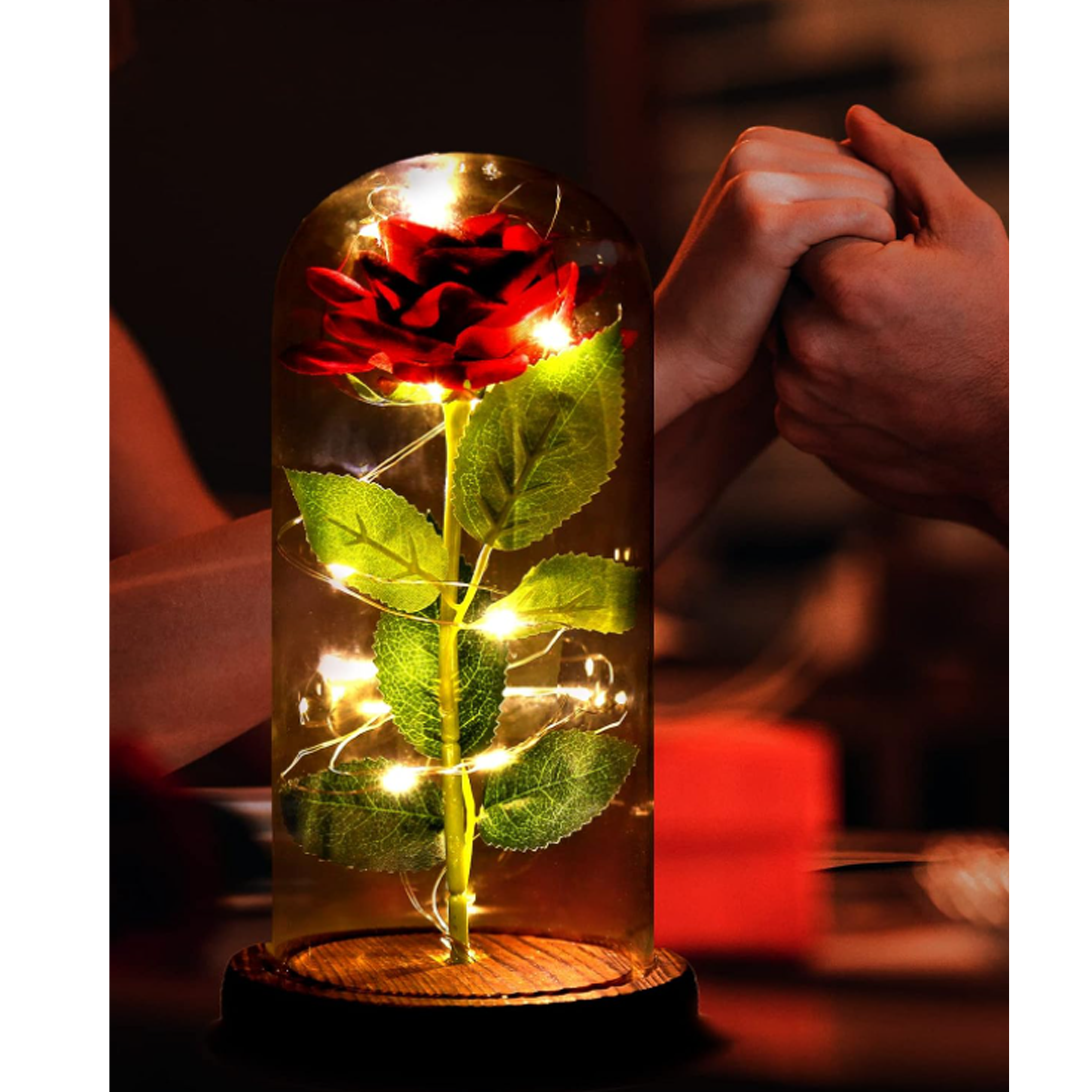 Eine Rose unter einer Glaskuppel mit Lichterketten, auf Holzsockel, mit Händen dahinter.