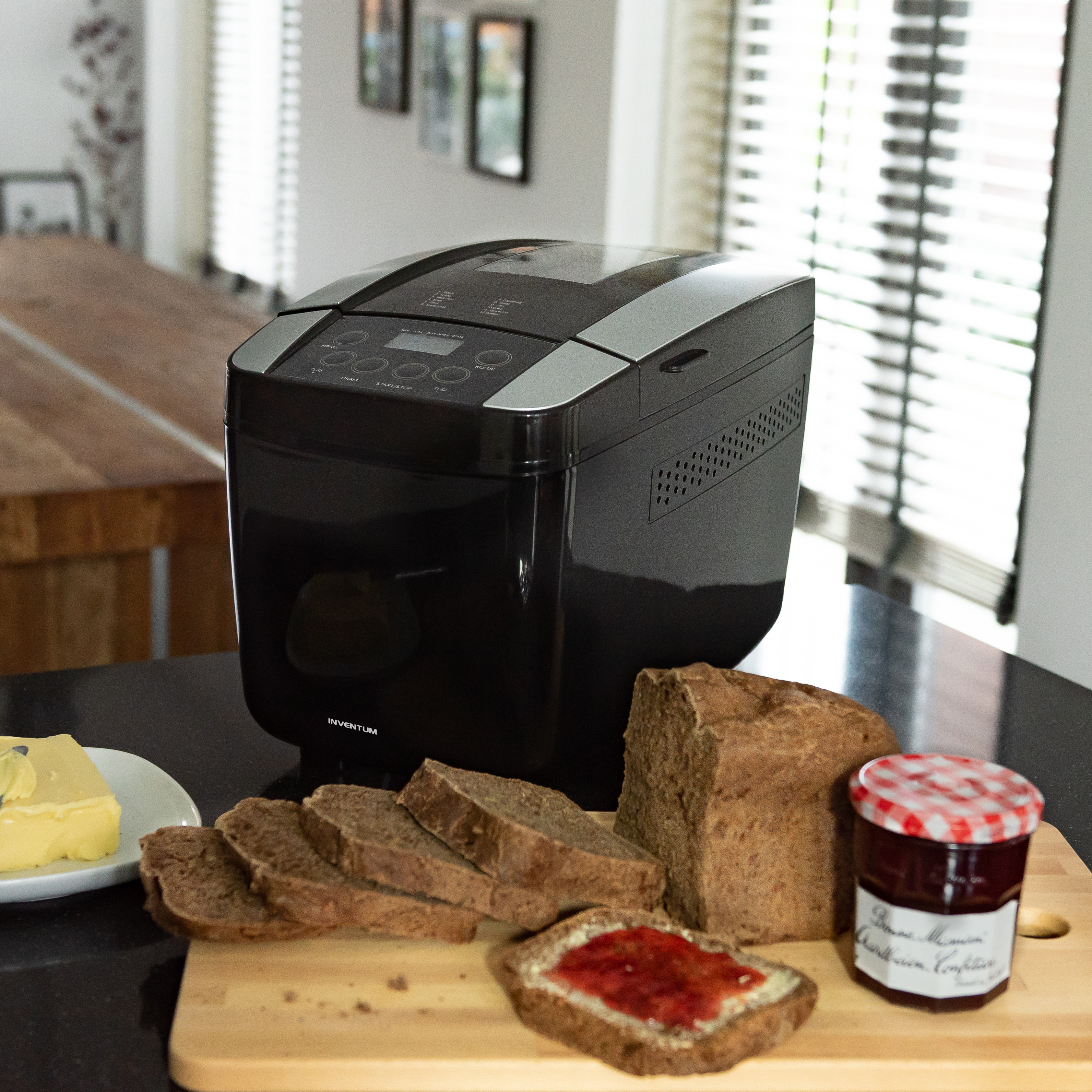 Black bread maker with bread, butter, jam, and sliced bread on a wooden board. Kitchen setting.