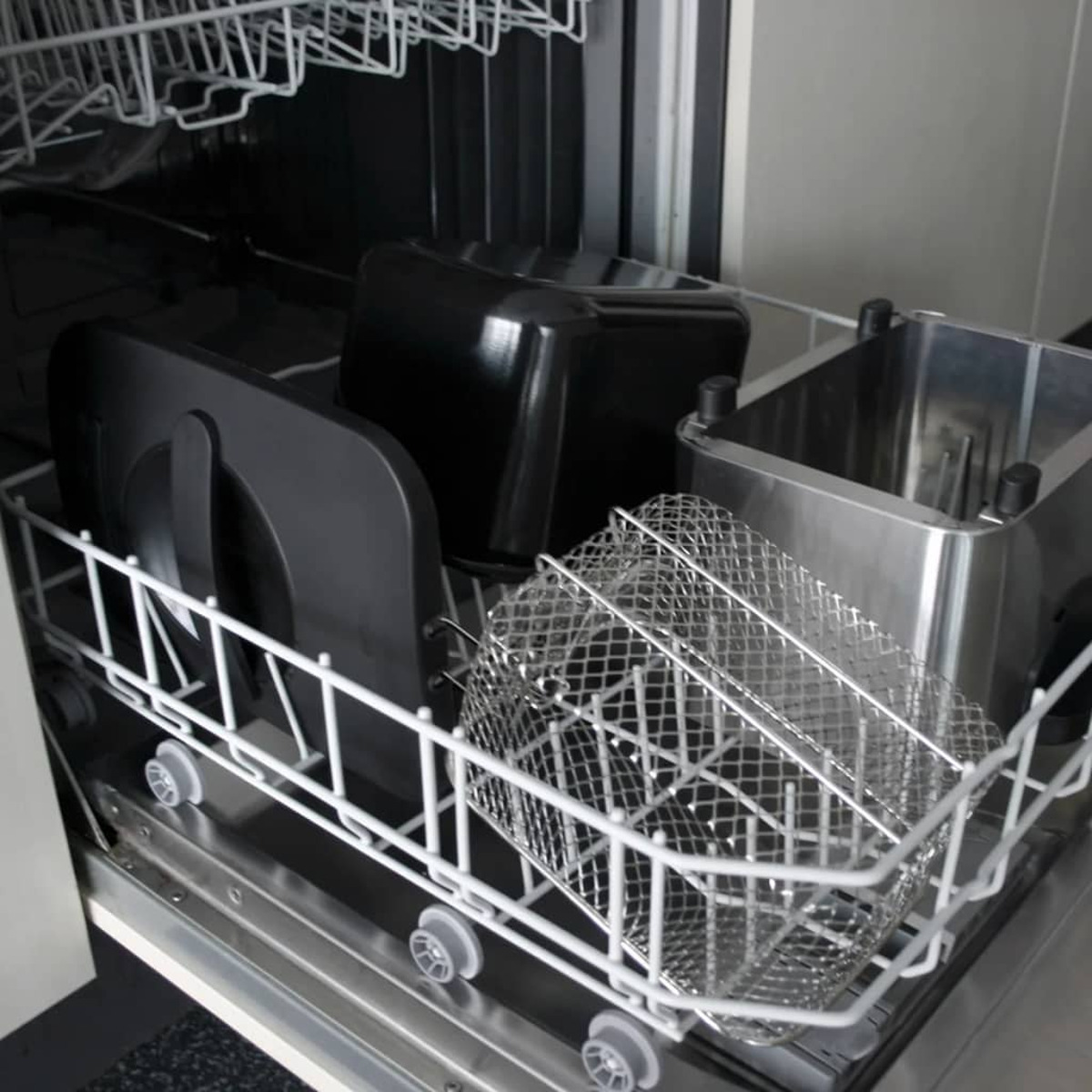 Vaatwasser met airfryer-onderdelen in de rekken. Bevat een draadmand, zwarte onderdelen en een metalen bak.