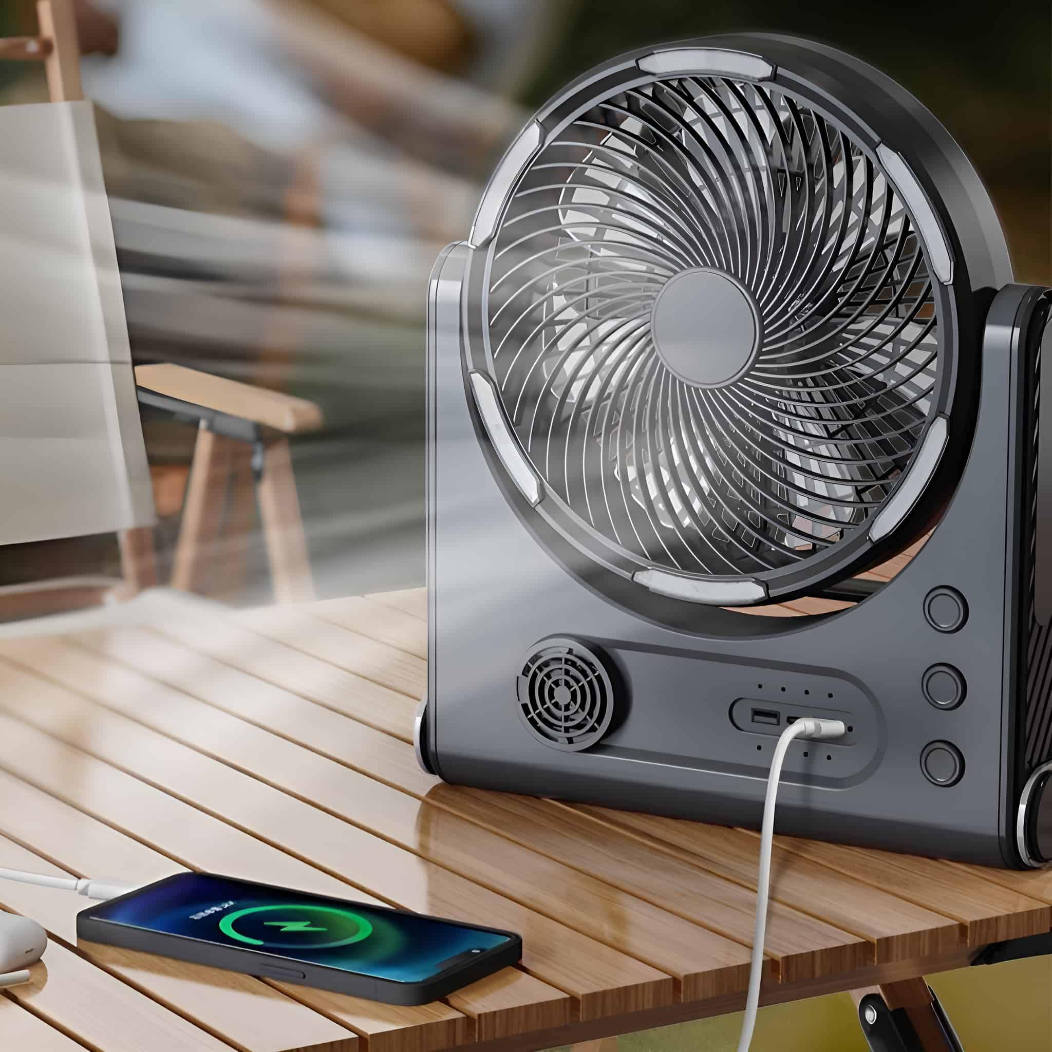 Schwarzer und grauer Ventilator auf einem Holztisch, der ein Telefon auflädt. Ein Stuhl im Hintergrund.