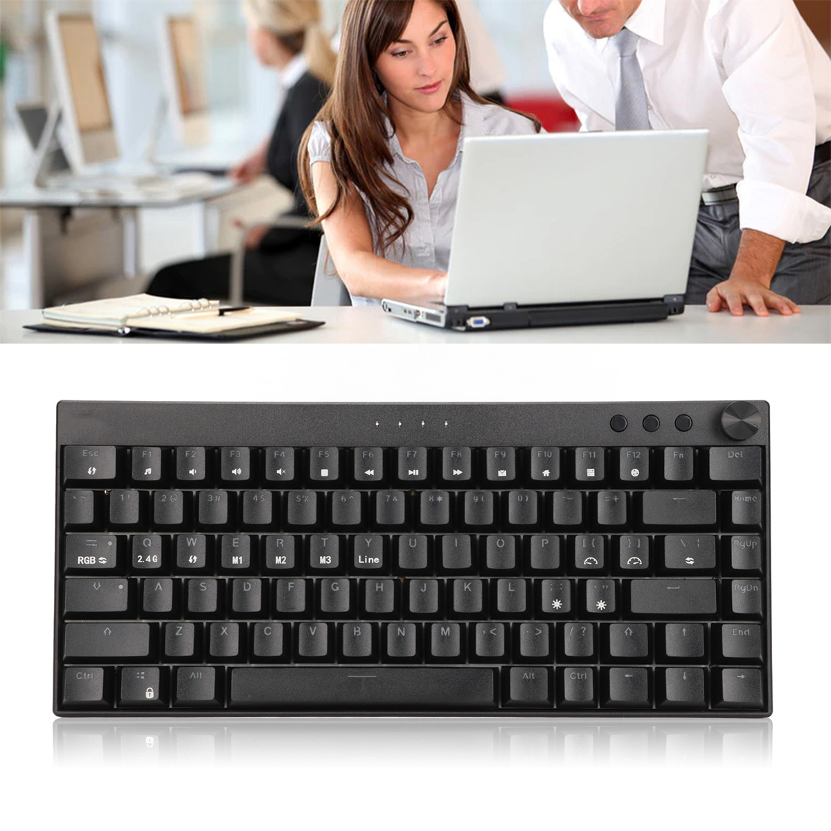 Eine schwarze Tastatur steht auf einem Schreibtisch, im Hintergrund ist ein Laptop zu sehen. Zwei Personen schauen auf den Laptop.