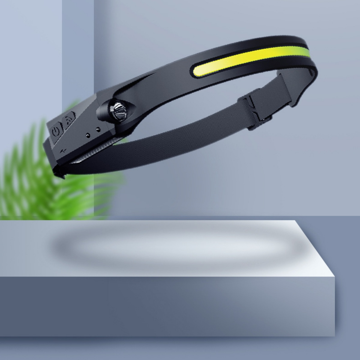 Eine schwarze Stirnlampe mit gelbem Lichtbalken, auf einer weißen Plattform, mit einer grünen Pflanze in der Ecke.