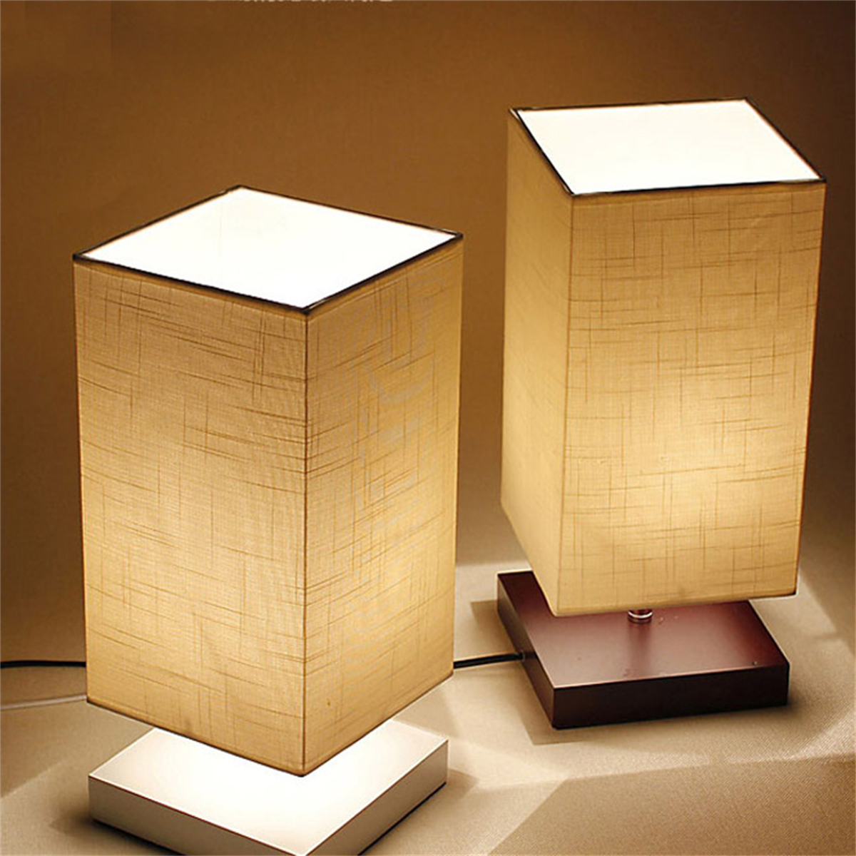 Zwei quadratische Lampen mit Stoffschirmen. Sie haben weiße Sockel und stehen auf einer dunklen Oberfläche, warm beleuchtet.