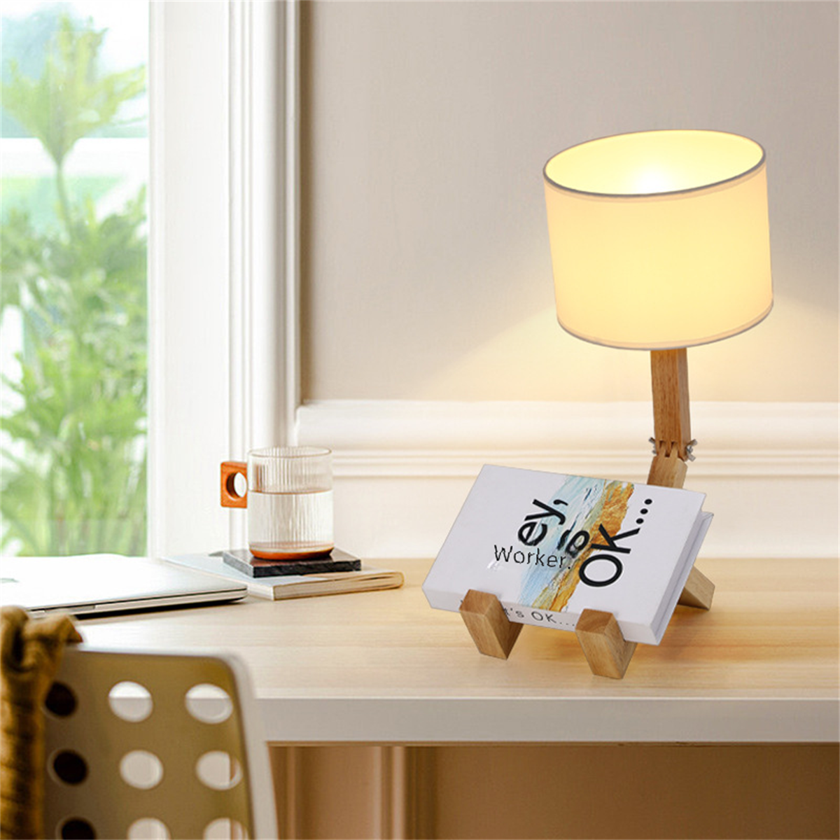 Eine Tischlampe mit Buchständer. Die Lampe hat einen weißen Schirm. Eine Tasse und ein Laptop sind auf dem Tisch.