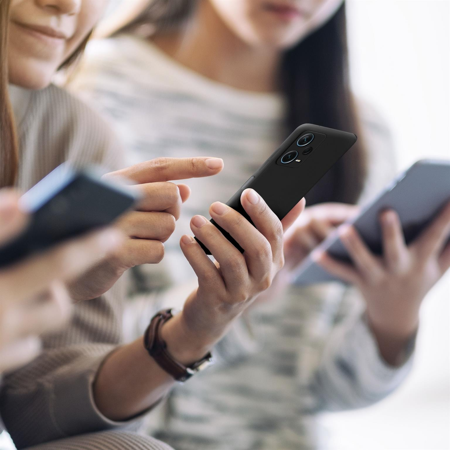 Drei Personen schauen auf Handys. Eine Person hält ein schwarzes Handy, die anderen schauen auch.