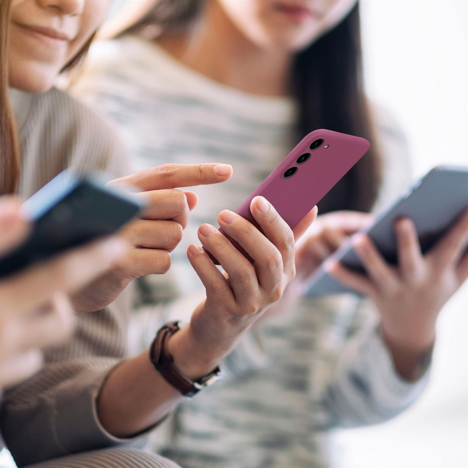 Drei Personen halten Telefone. Eine Hand zeigt auf ein lila Telefon. Der Hintergrund ist unscharf.