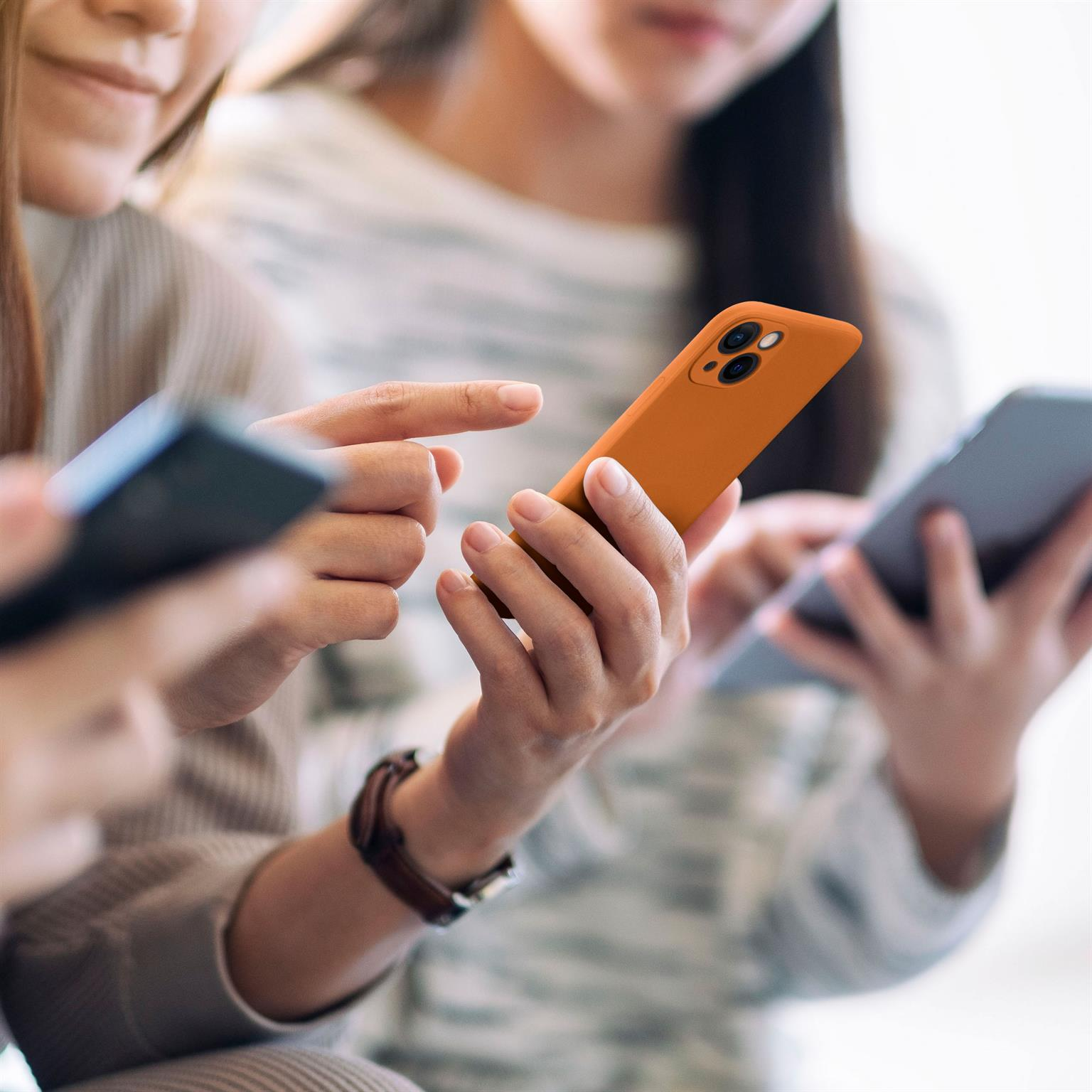 Drei Personen halten Telefone; eines orange, zwei schwarz, alle schauen auf Bildschirme.