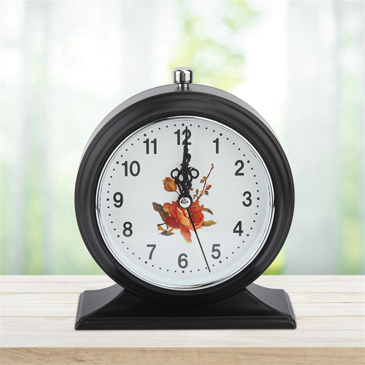 Schwarze Uhr mit floralem Zifferblatt, Zeiger auf 12:30. Schwarzer Sockel, Holztisch, Hintergrund unscharf.