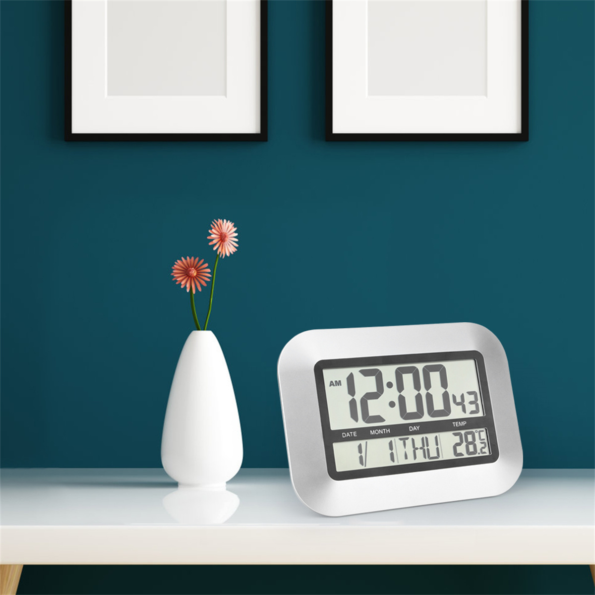 Eine Digitaluhr mit weißen Zahlen. Zwei gerahmte Bilder sind darüber. Eine Vase mit Blumen befindet sich links.
