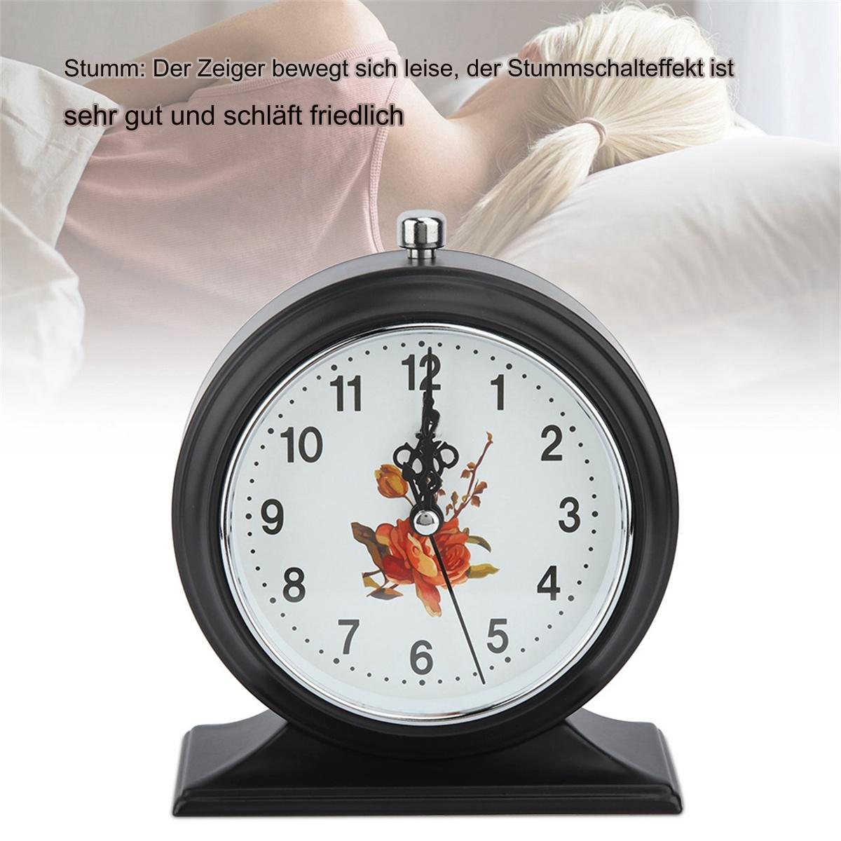 Ein schwarzer Wecker mit floralem Design, im Hintergrund schläft eine Person.