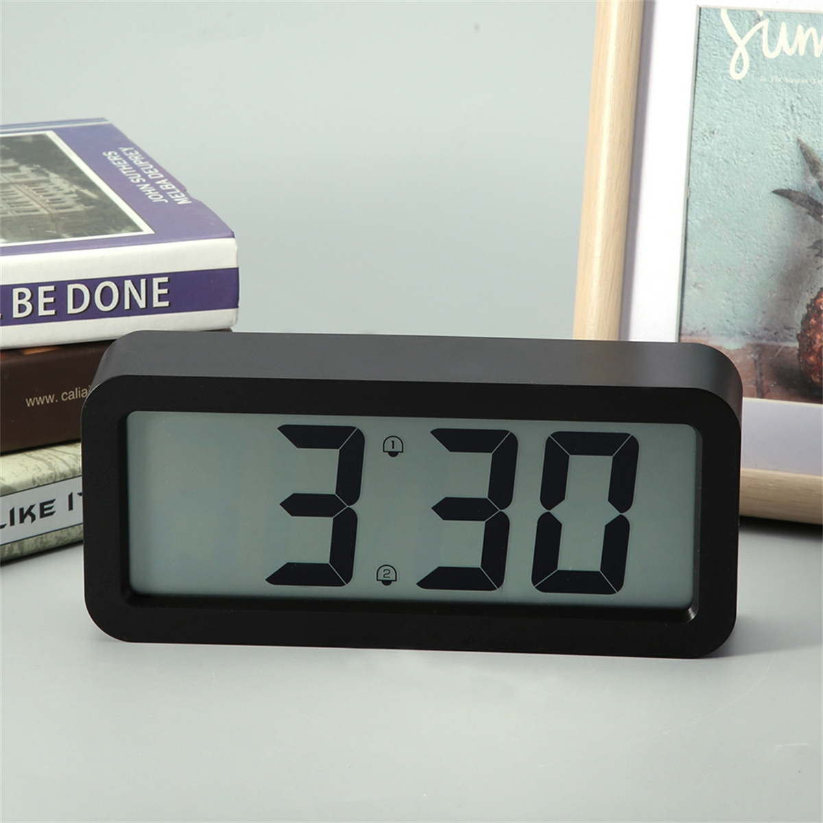 Eine schwarze Digitaluhr, die 3:30 anzeigt. Sie steht auf einem Holztisch mit Büchern und einem Rahmen im Hintergrund.