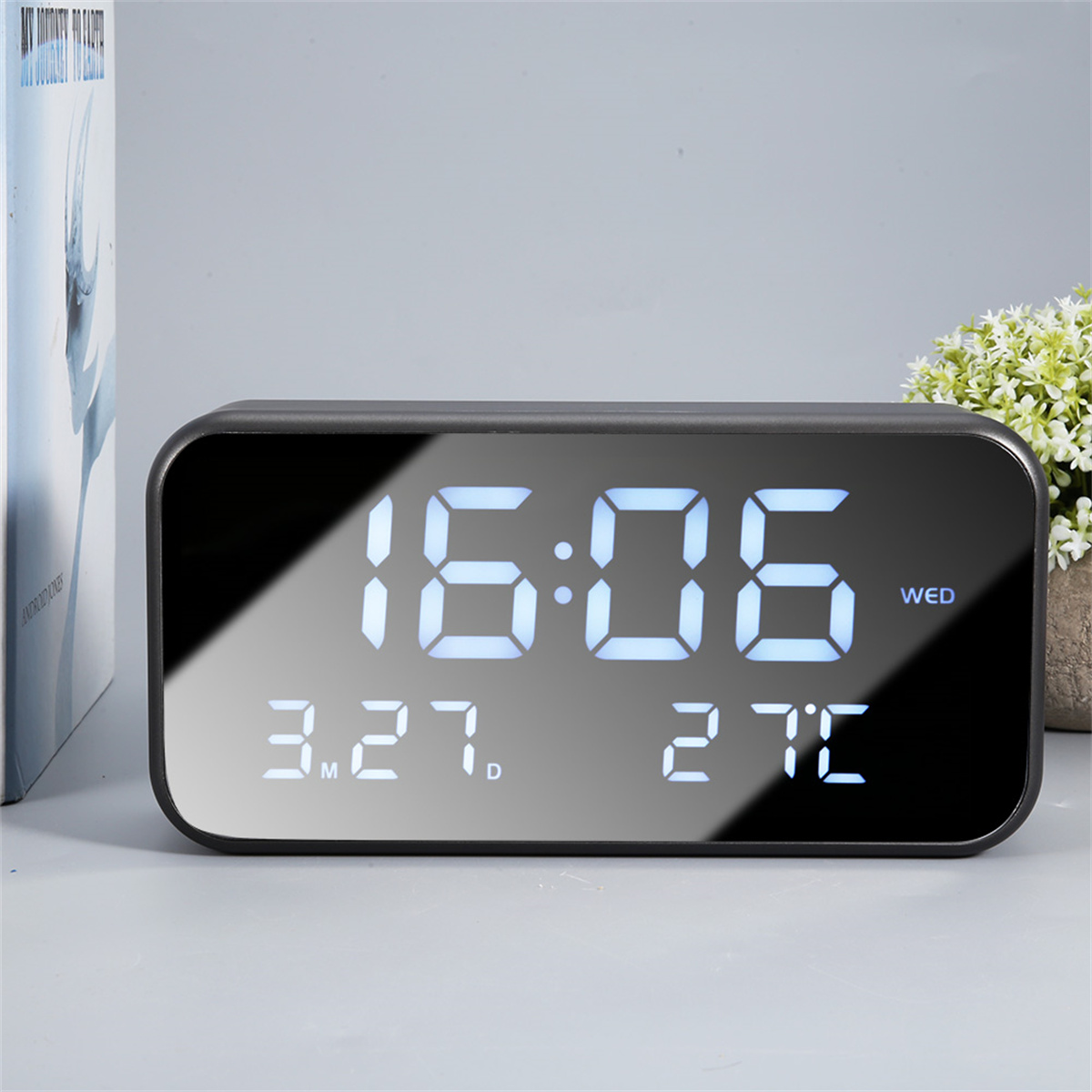 Schwarze Digitaluhr zeigt 16:06 mit Datum und Temperatur. Buch und Pflanze im Hintergrund.
