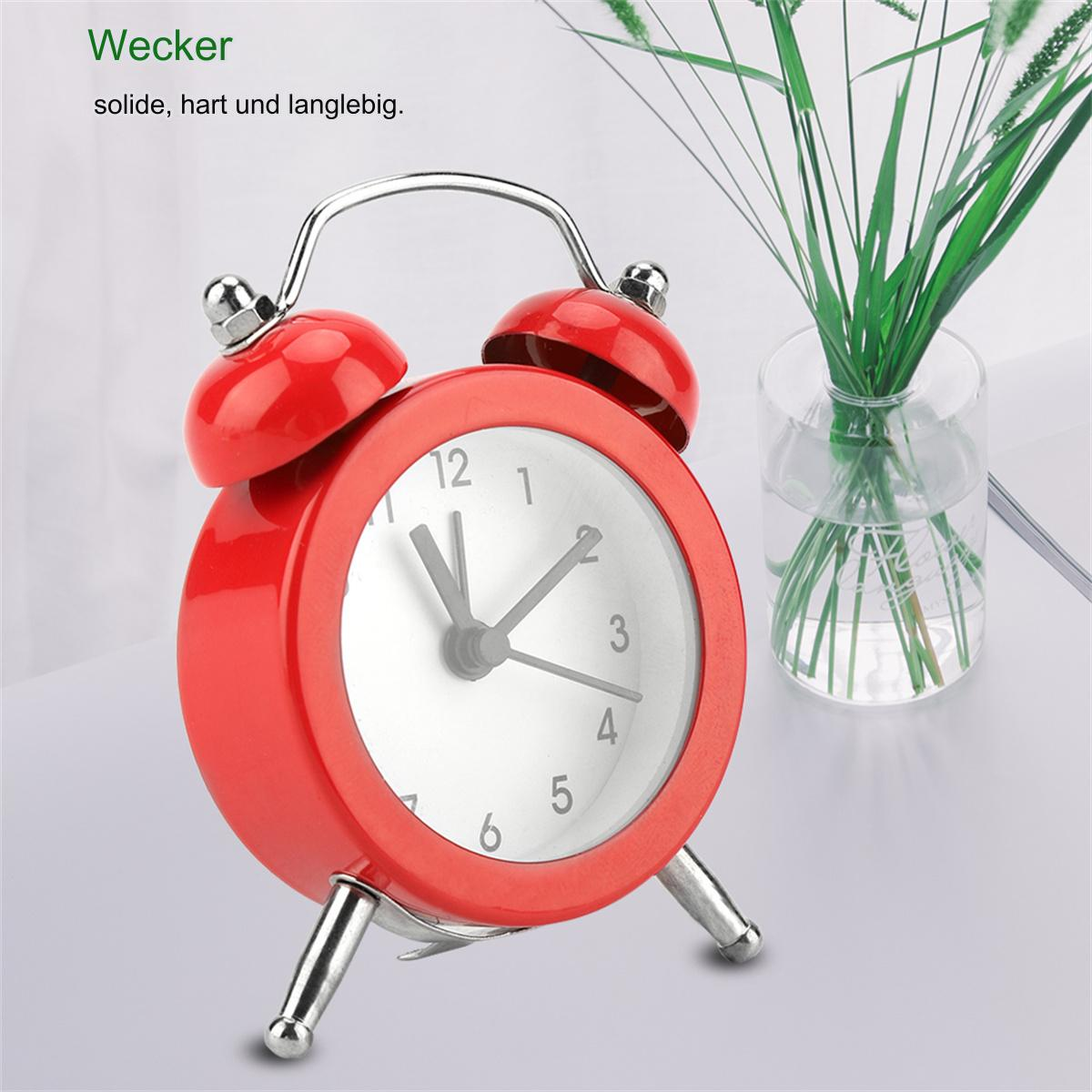 Ein roter Wecker mit weißem Zifferblatt, der die Uhrzeit anzeigt. Eine Vase mit grünem Gras ist im Hintergrund.
