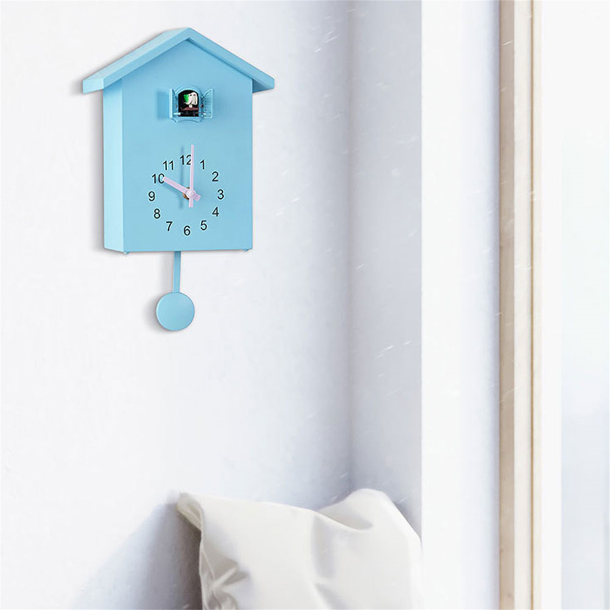 Blaue Kuckucksuhr an einer weißen Wand. Die Uhr zeigt die Zeit, 10:10. Ein weißes Kissen darunter.