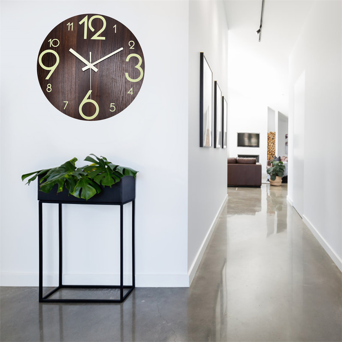 Eine Wanduhr mit römischen Ziffern. Eine Pflanze in einem schwarzen Topf steht darunter. Ein Flur ist im Hintergrund.