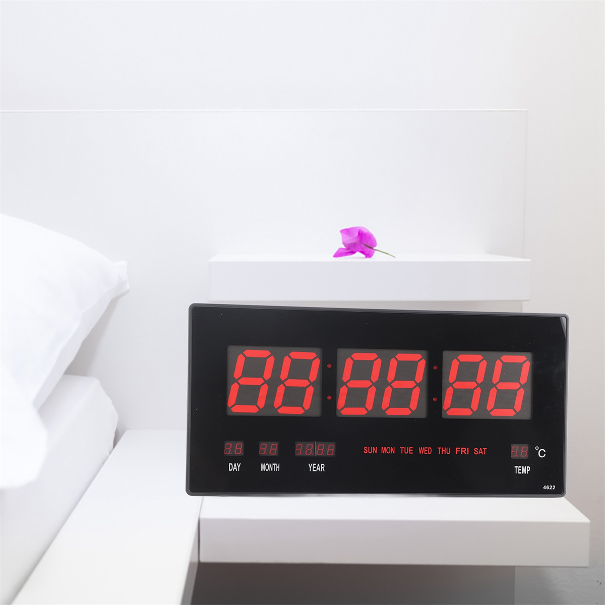Schwarze Digitaluhr mit roten Ziffern, zeigt Uhrzeit, Datum und Temperatur an. Lila Blume auf weißem Hintergrund.