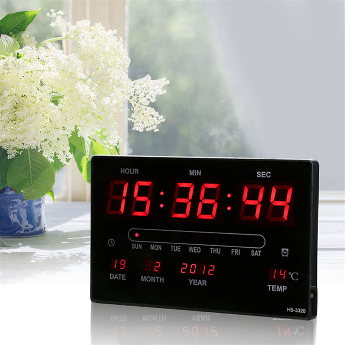 Schwarze Digitaluhr zeigt Zeit, Datum und Temperatur an. Weiße Blumen sind im Hintergrund.