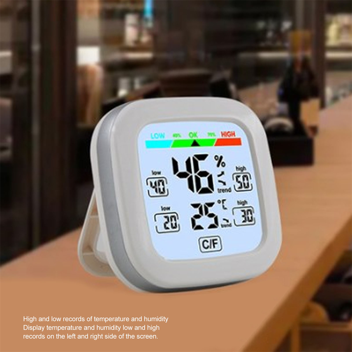Weißes digitales Thermometer zeigt Feuchtigkeits- und Temperaturwerte. Im Hintergrund ein Holztisch.