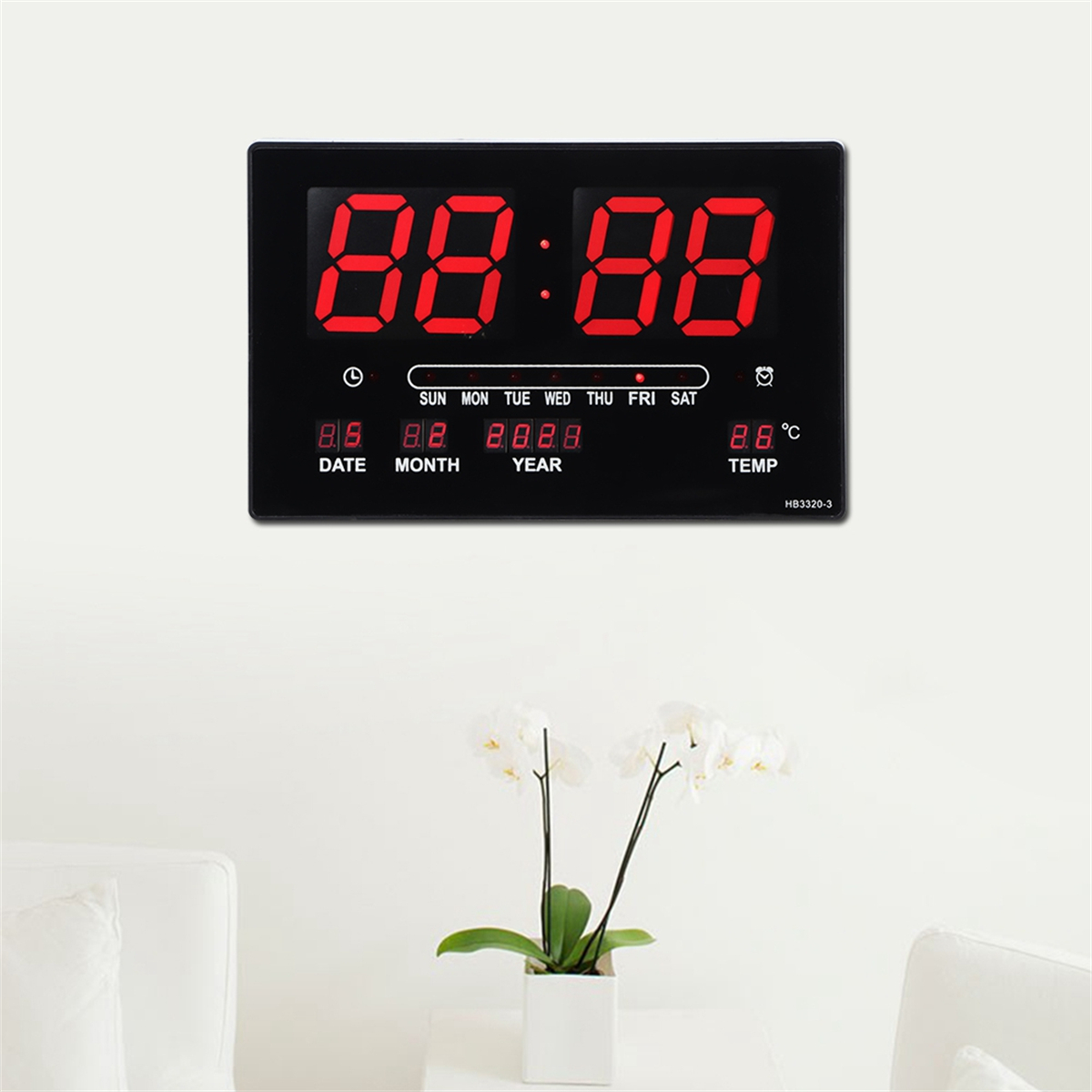 Schwarze Digitaluhr zeigt Zeit und Datum. Weißer Hintergrund, Blumen darunter.