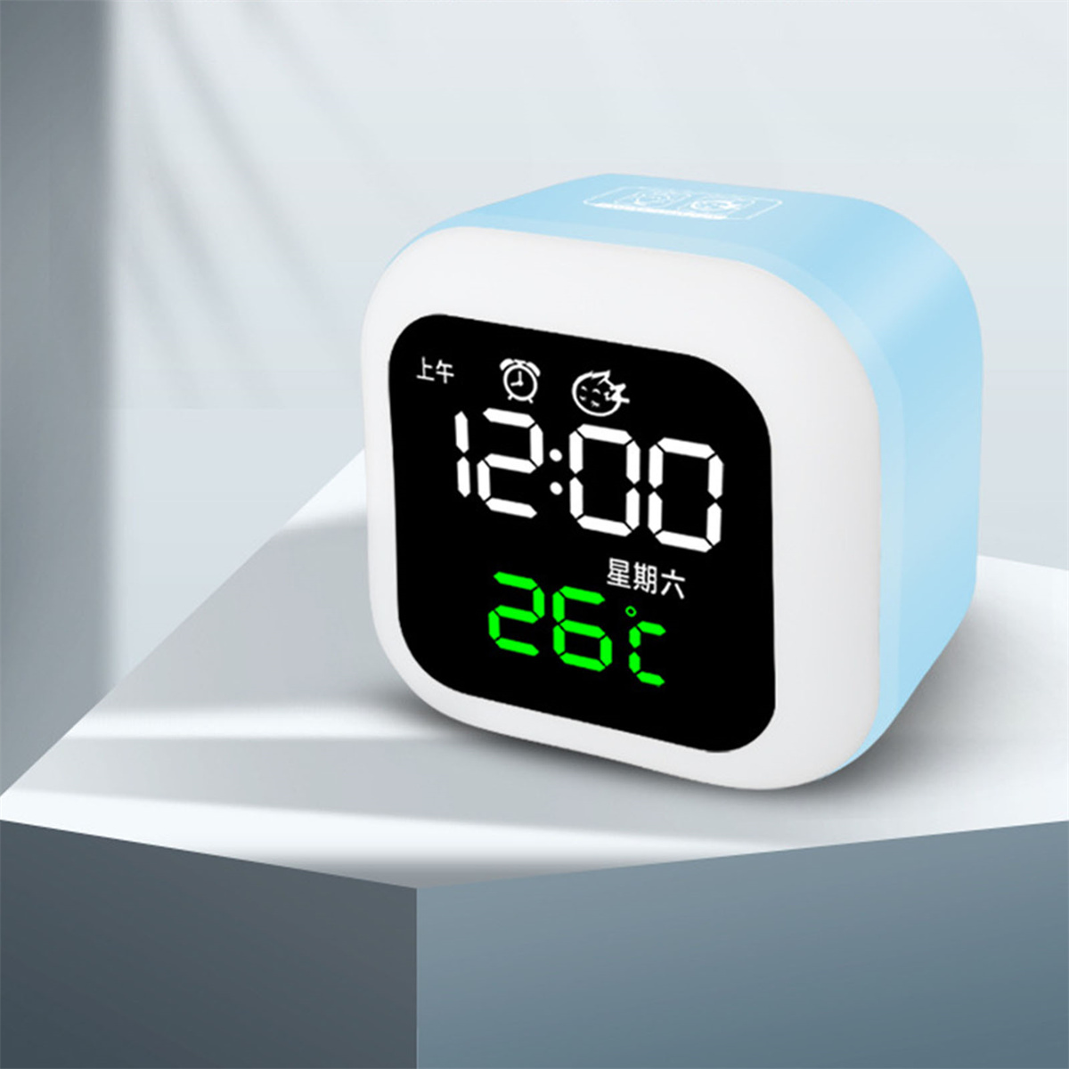 Eine hellblaue Digitaluhr, die 12:00 und 26°C anzeigt. Es steht auf einer weißen Oberfläche mit weißem Hintergrund.
