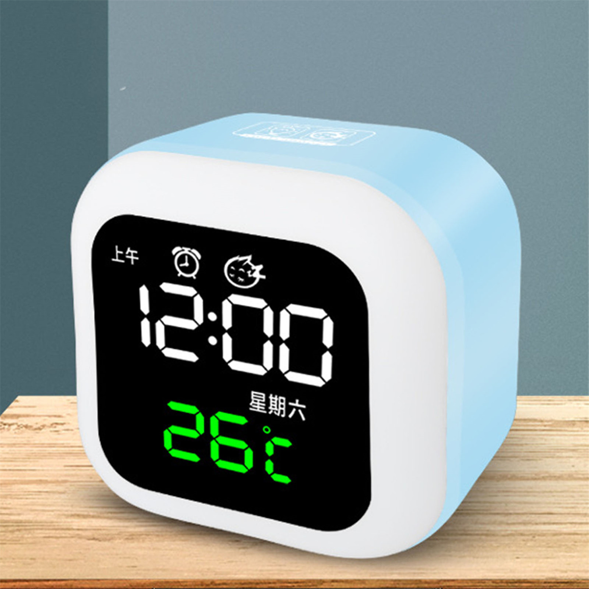 Eine blau-weiße Digitaluhr, die 12:00 und 26°C anzeigt. Die Uhr steht auf einer Holzoberfläche.