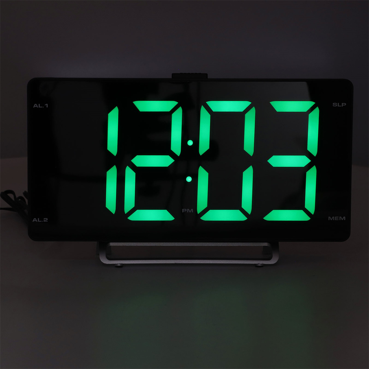 Digitaluhr mit 12:03 PM mit grünen Zahlen. Schwarzes Gehäuse, auf grauer Oberfläche. Dunkler Hintergrund.