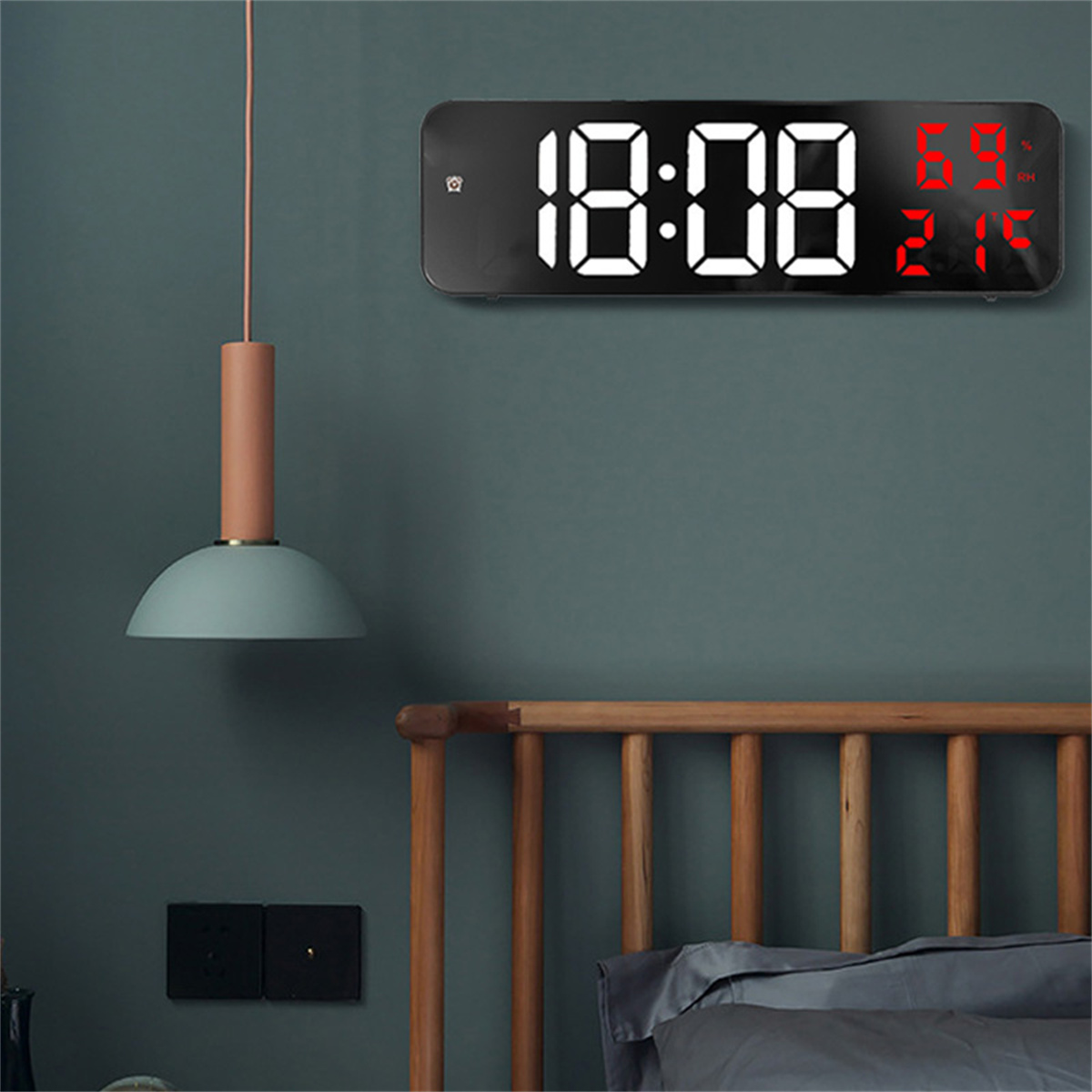 Eine Digitaluhr zeigt 18:08. Sie hat Feuchtigkeits- und Temperaturmesswerte. Über einem Bett hängt eine Lampe.