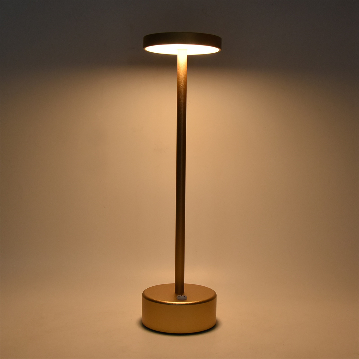 Goldfarbene hohe Lampe, oben beleuchtet, auf goldenem Sockel vor weißem Hintergrund.