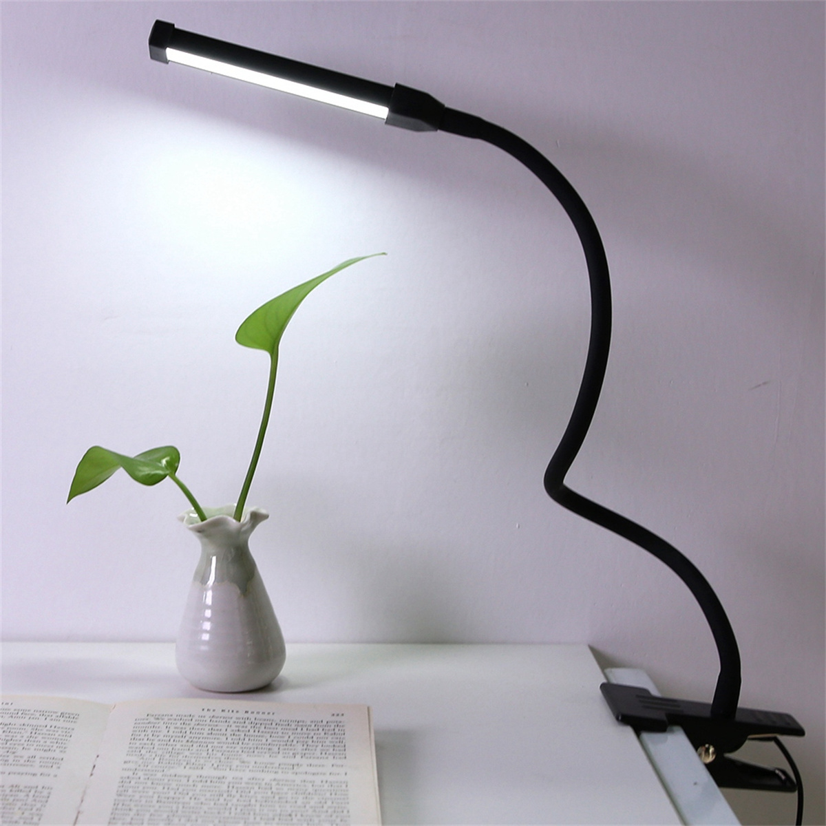 Eine schwarze, flexible Schreibtischlampe mit einer kleinen Vase mit einer Pflanze. Das Licht ist an.