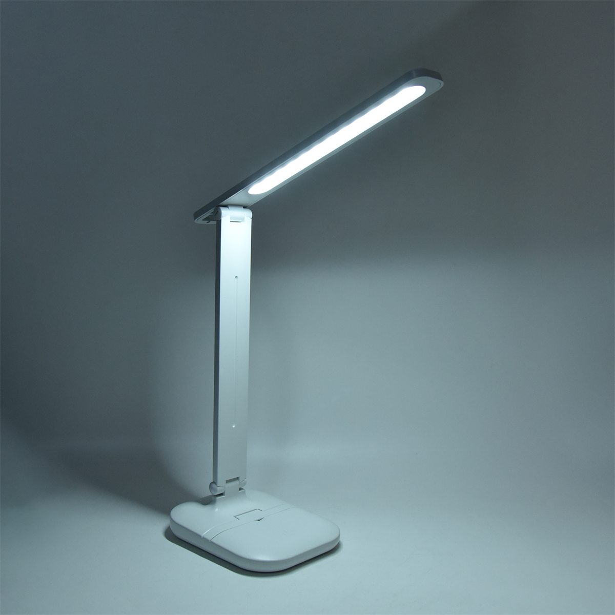Eine weiße Schreibtischlampe mit LED-Licht leuchtet. Sie steht auf einer weißen Oberfläche vor grauem Hintergrund.